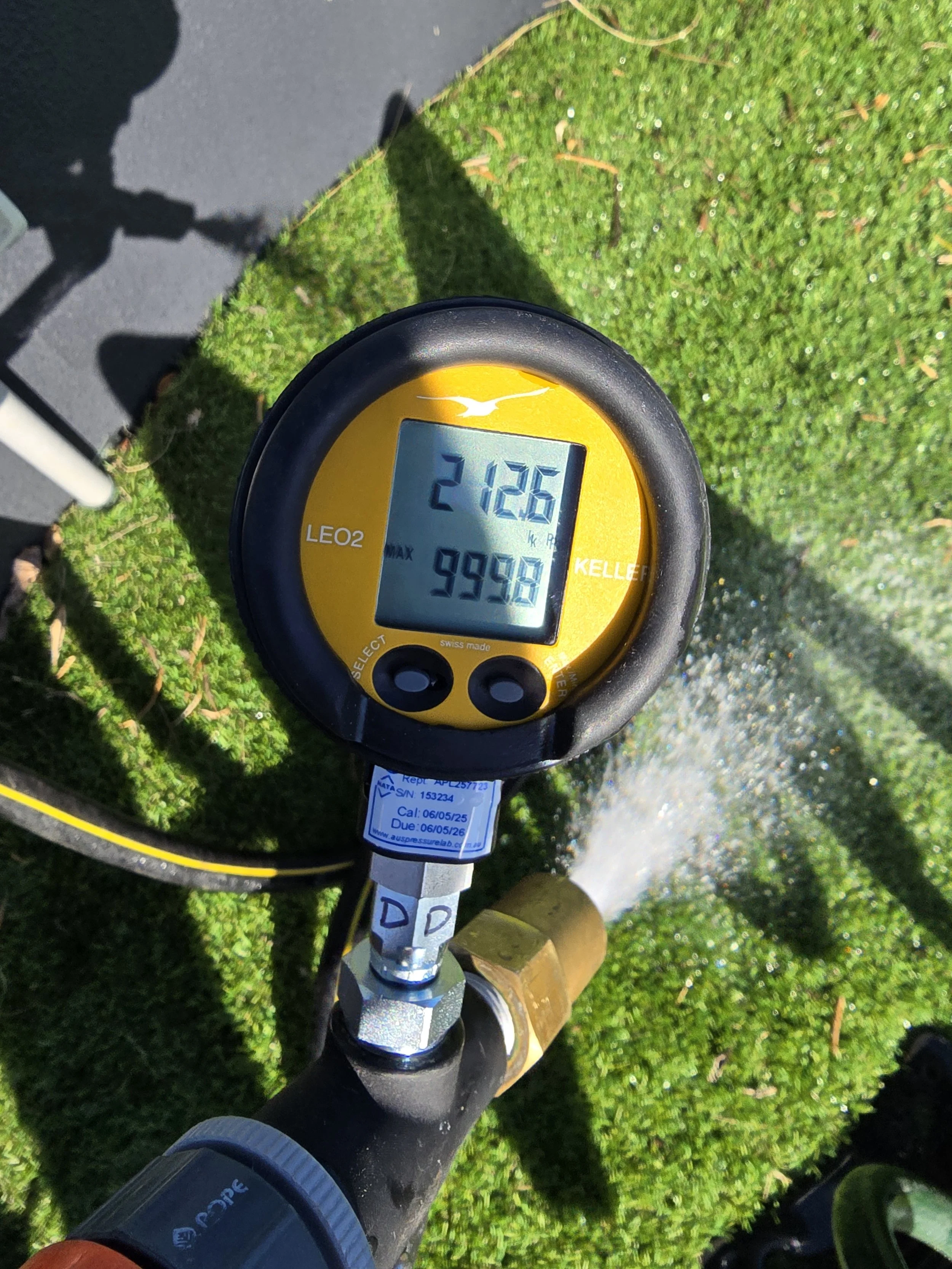 A digital water meter showing readings for AAMA 501.2 water hose testing, spraying water onto a patch of green grass.