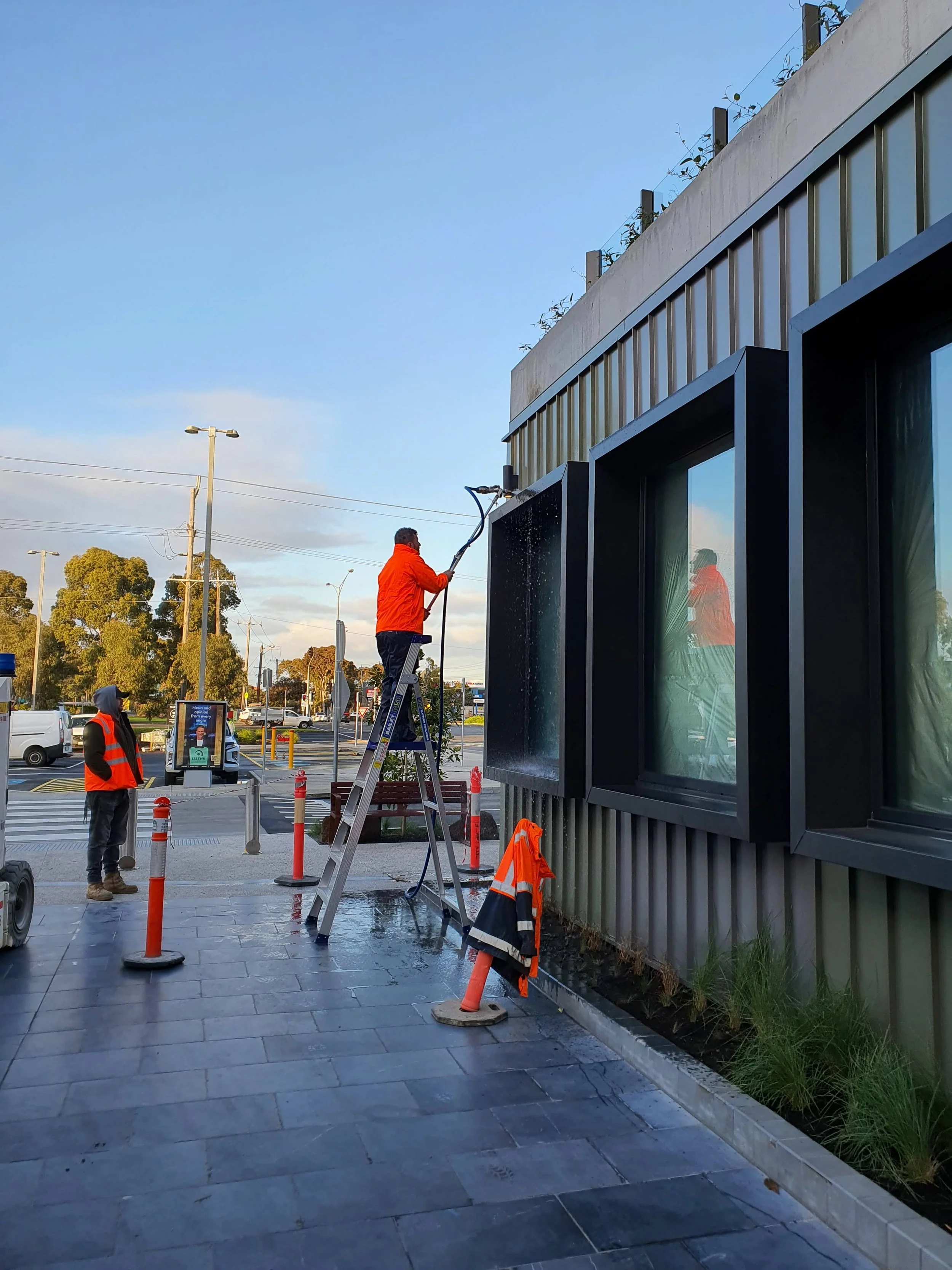 Facade engineer performing a AAMA 501.2 test on the exterior window of a building using a water pole and hose, with another technician consulting nearby, on a metropolitan sidewalk with traffic and trees in the background.
