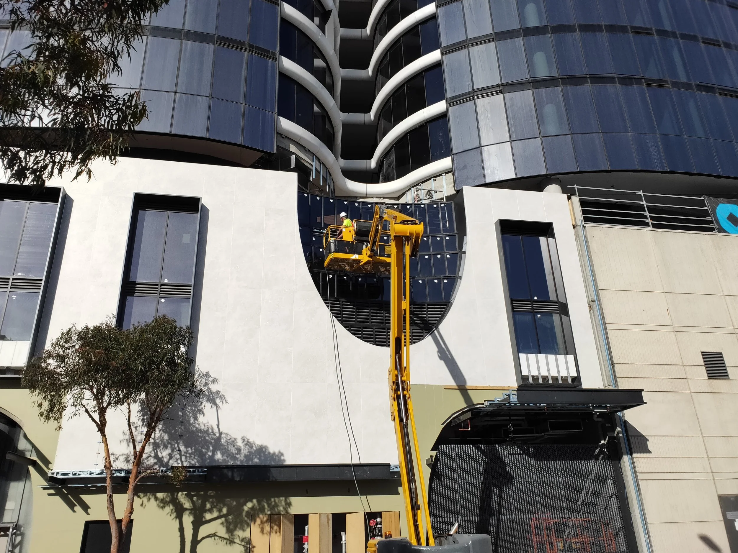 Facade testers on a yellow boom lift testing the facade of a modern building with unique, curved black glass windows on a construction site in the city.