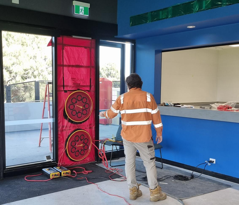 Facade technician in an orange safety shirt and beige pants standing inside a building near a large window performing blower door testing, with airtightness testing equipment and tools around.