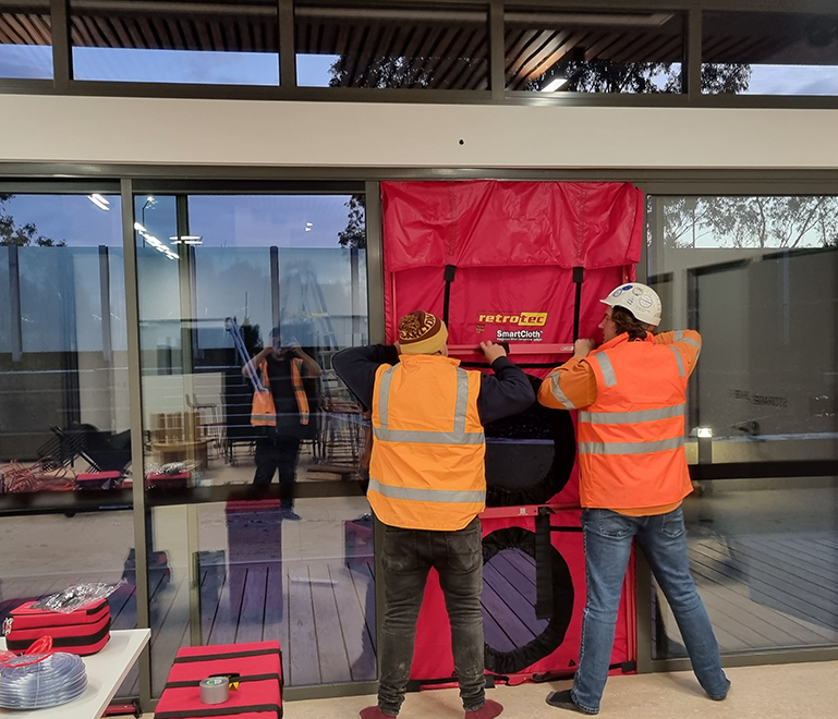 Two building envelope engineers install a red RetroTec SmartCloth weatherproof barrier on the open door of a building for blowerdoor testing. They are wearing orange safety vests and helmets, facing away from the camera.