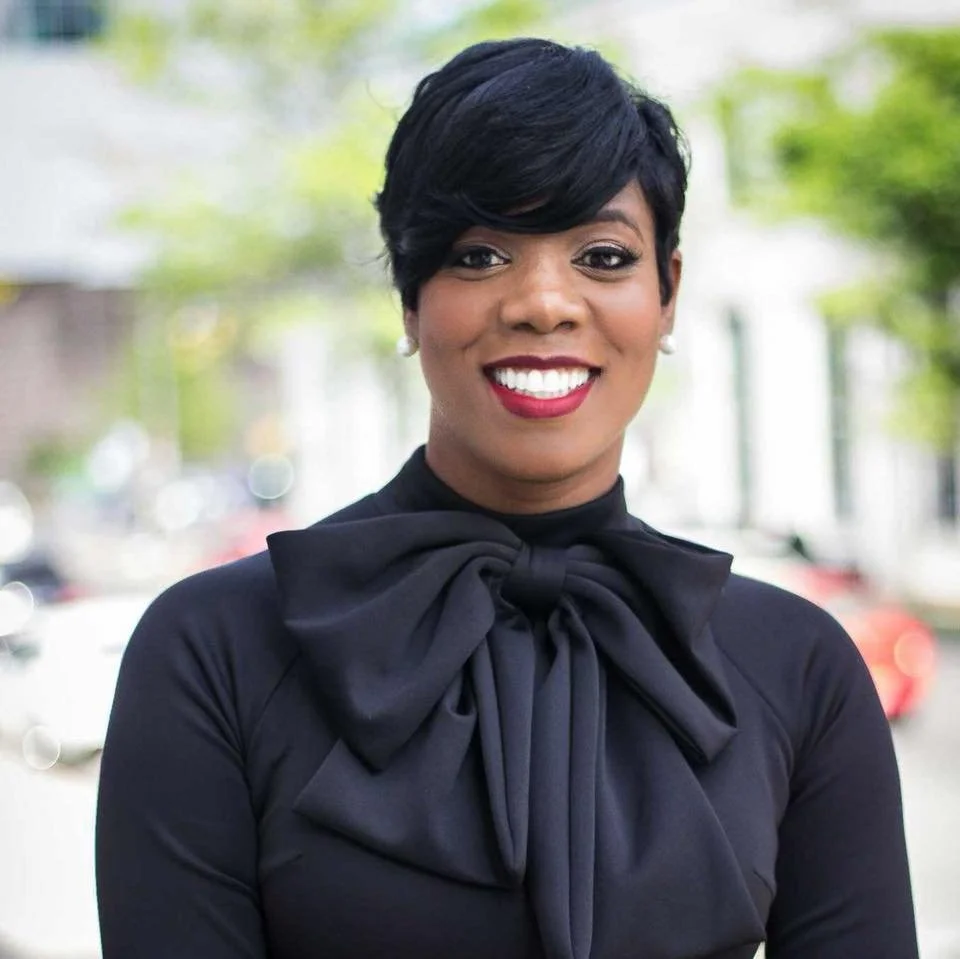 A woman with short black hair, wearing a black top with a large bow, smiling outdoors with blurred greenery and cars in the background.