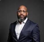 A man with glasses, a beard, and a bald head, wearing a dark suit and white shirt, posing against a gray background.