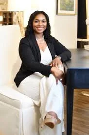 A woman with dark hair wearing a black blazer and white pants, sitting on a white sofa in an indoor setting with artwork on the wall behind her.