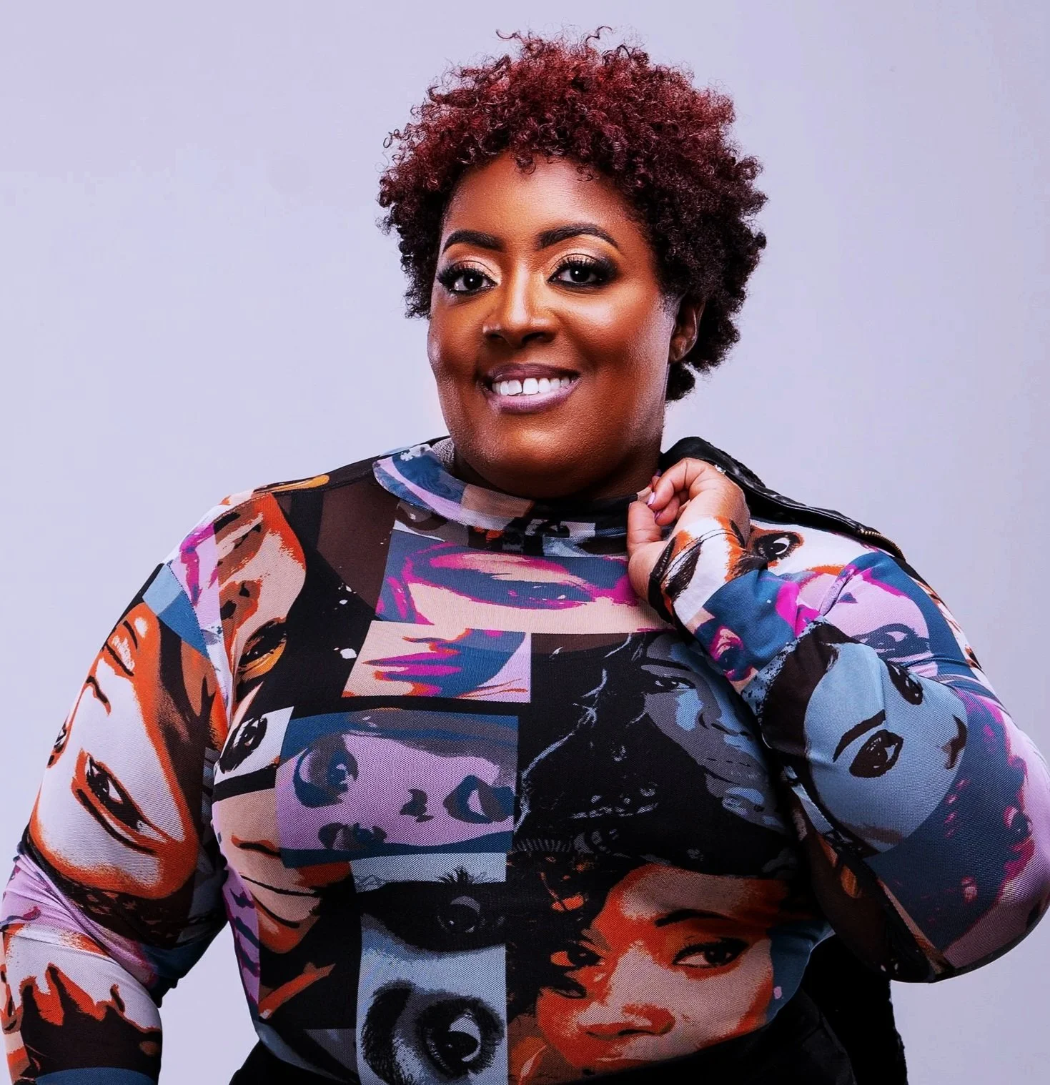 A woman with short, curly hair wearing a colorful, patterned top featuring various faces and artwork, posing against a plain gray background.