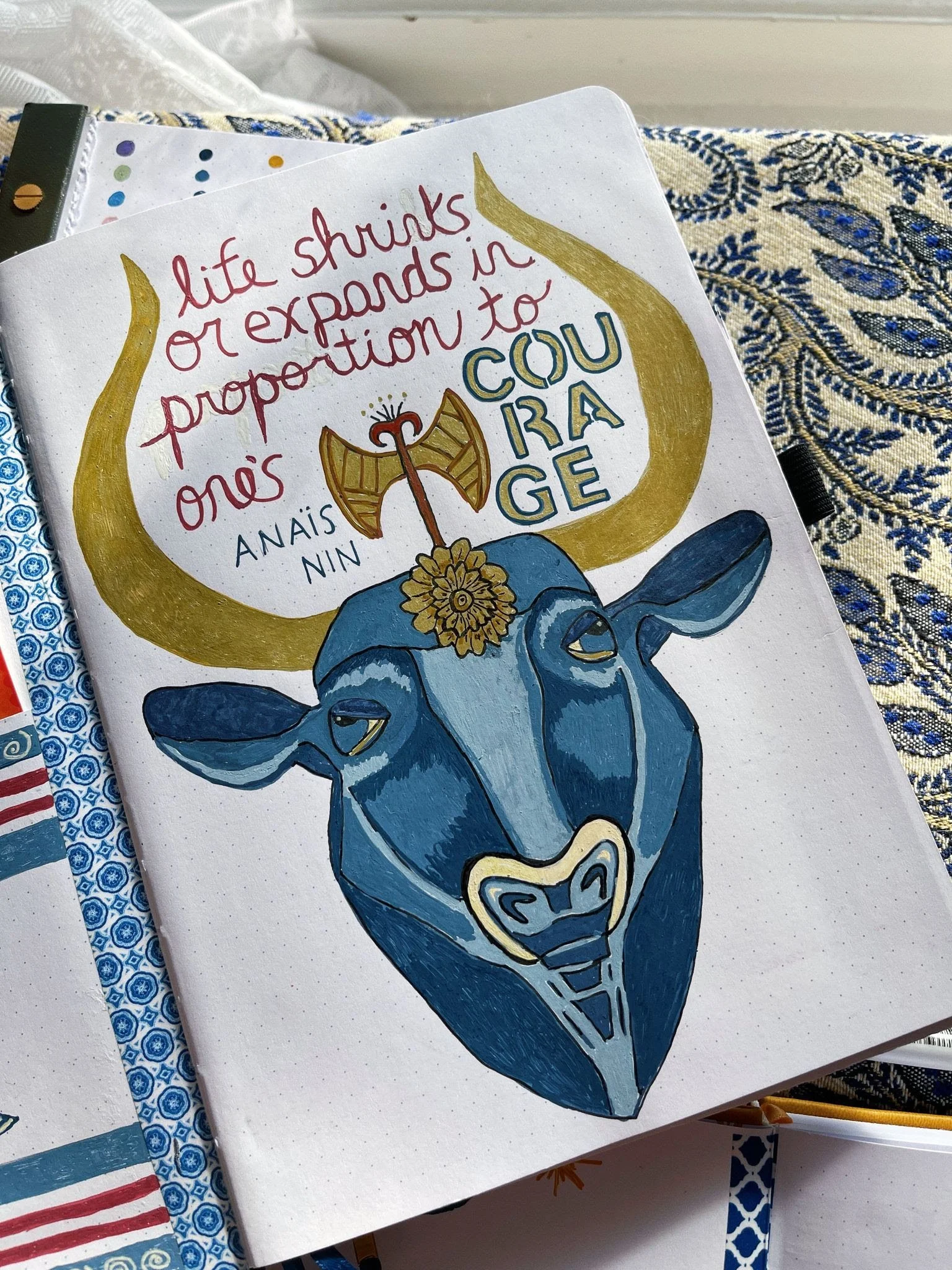 An illustration of a Minotaur's head with large horns, with the horns forming a quote saying, 'life shrinks or expands in proportion to one's courage,' written inside the horns. The quote is in red and blue handwriting.