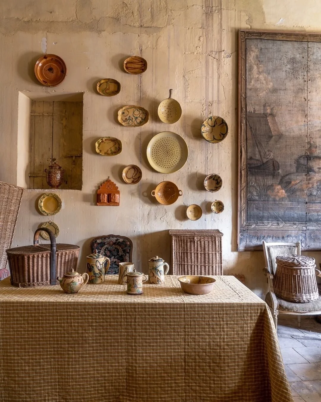 A rustic dining room with a beige checkered tablecloth on a wooden table, decorated with ceramic pitchers and bowls. A wicker basket and aged wooden chairs are around the table. The wall behind has a display of various clay and ceramic bowls, plates,