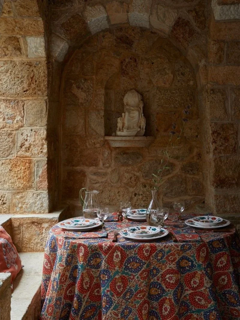 A round table decorated with a colorful, patterned tablecloth set with four white plates, napkins, glasses, a glass pitcher, and a tall glass vase with a single branch, inside a stone-walled room with a small statue in a niche.