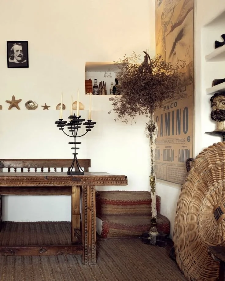 Decorative interior corner with a wooden table, a black candelabrum, a wall shelf with shells and starfish, a framed black-and-white portrait, a vintage poster, a potted shrub, and woven furniture.