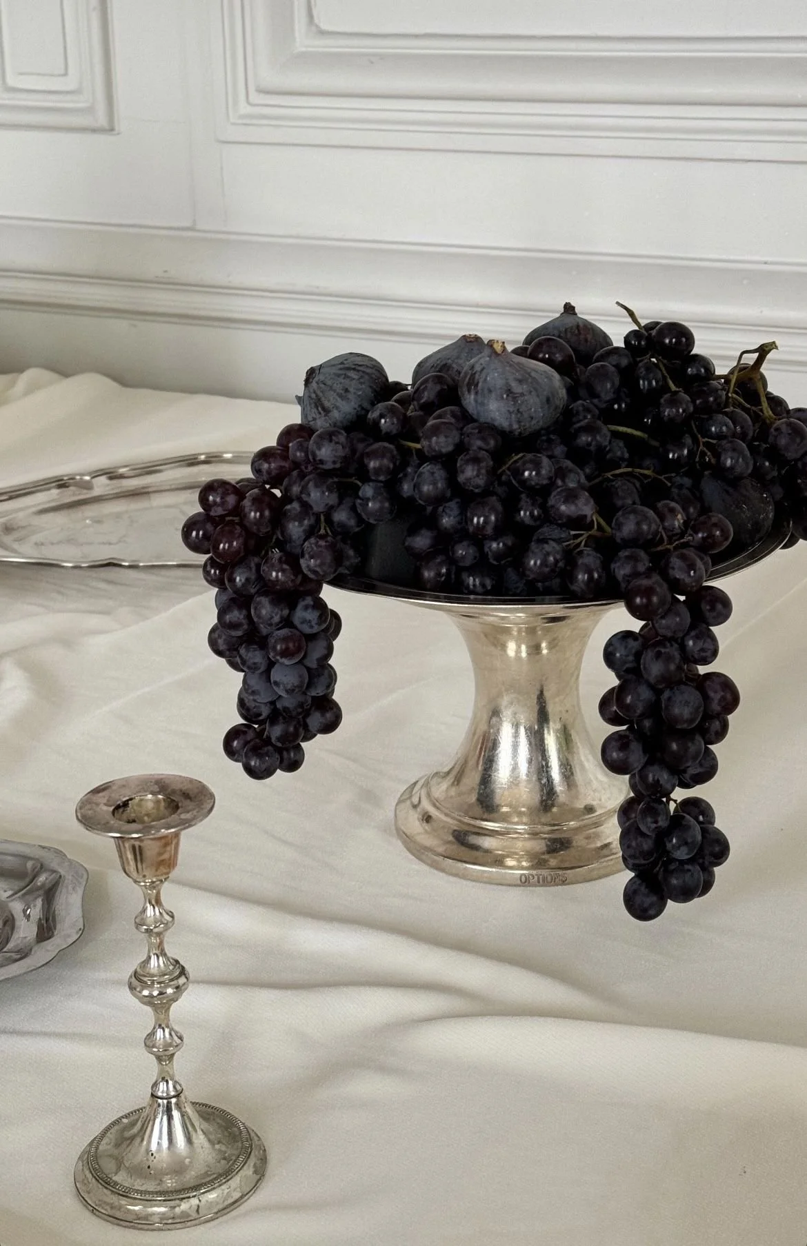 A silver pedestal bowl filled with black grapes and large dark figs on display on a white tablecloth.