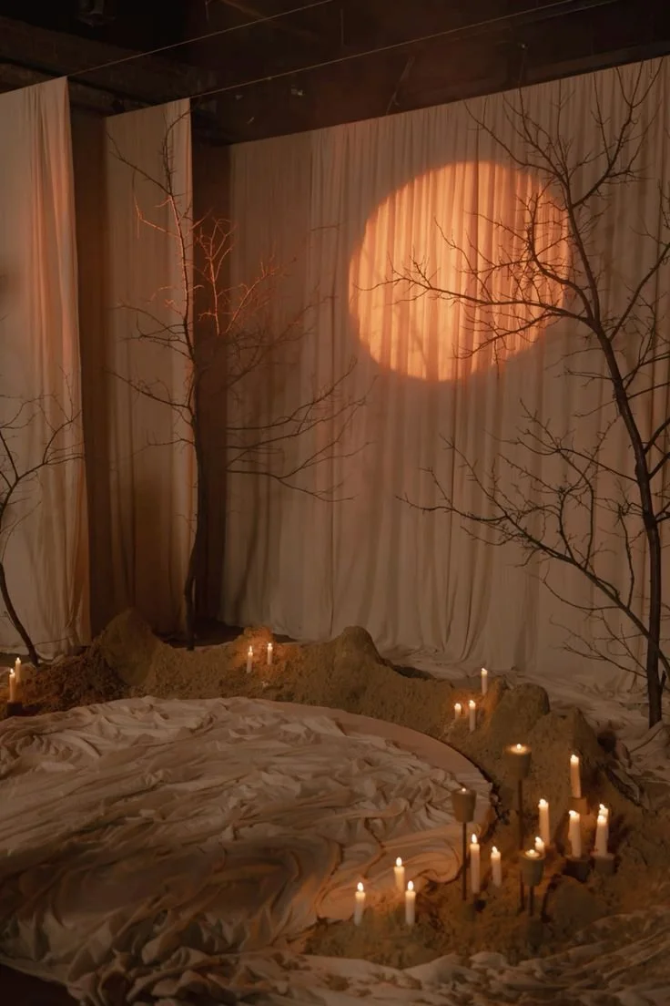 A whimsical indoor scene with a circular bed surrounded by candles, two leafless trees, and a beige curtain backdrop illuminated by a moon-shaped light projection.
