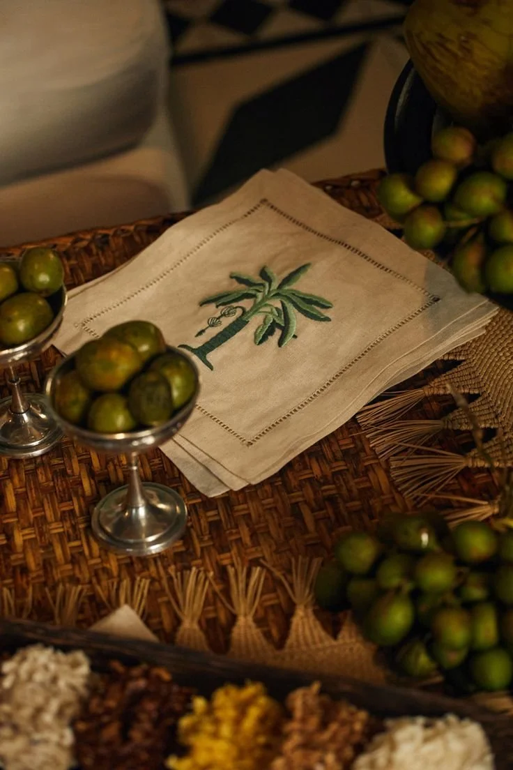 Embroidery of a palm tree on a cloth napkin on a woven table, surrounded by small green limes and other fruit.