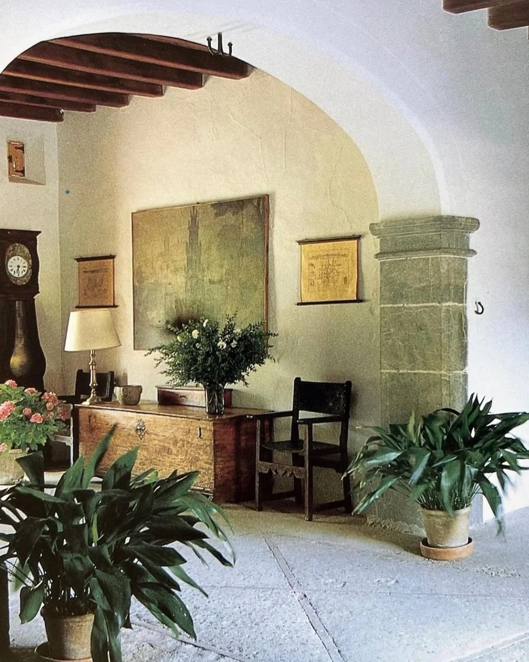 A cozy corner of a living room featuring a wooden chest with a large bouquet of white flowers, potted green plants, a vintage wall clock, framed artwork, a lamp, and a black chair, with an arched ceiling and stone pillar.