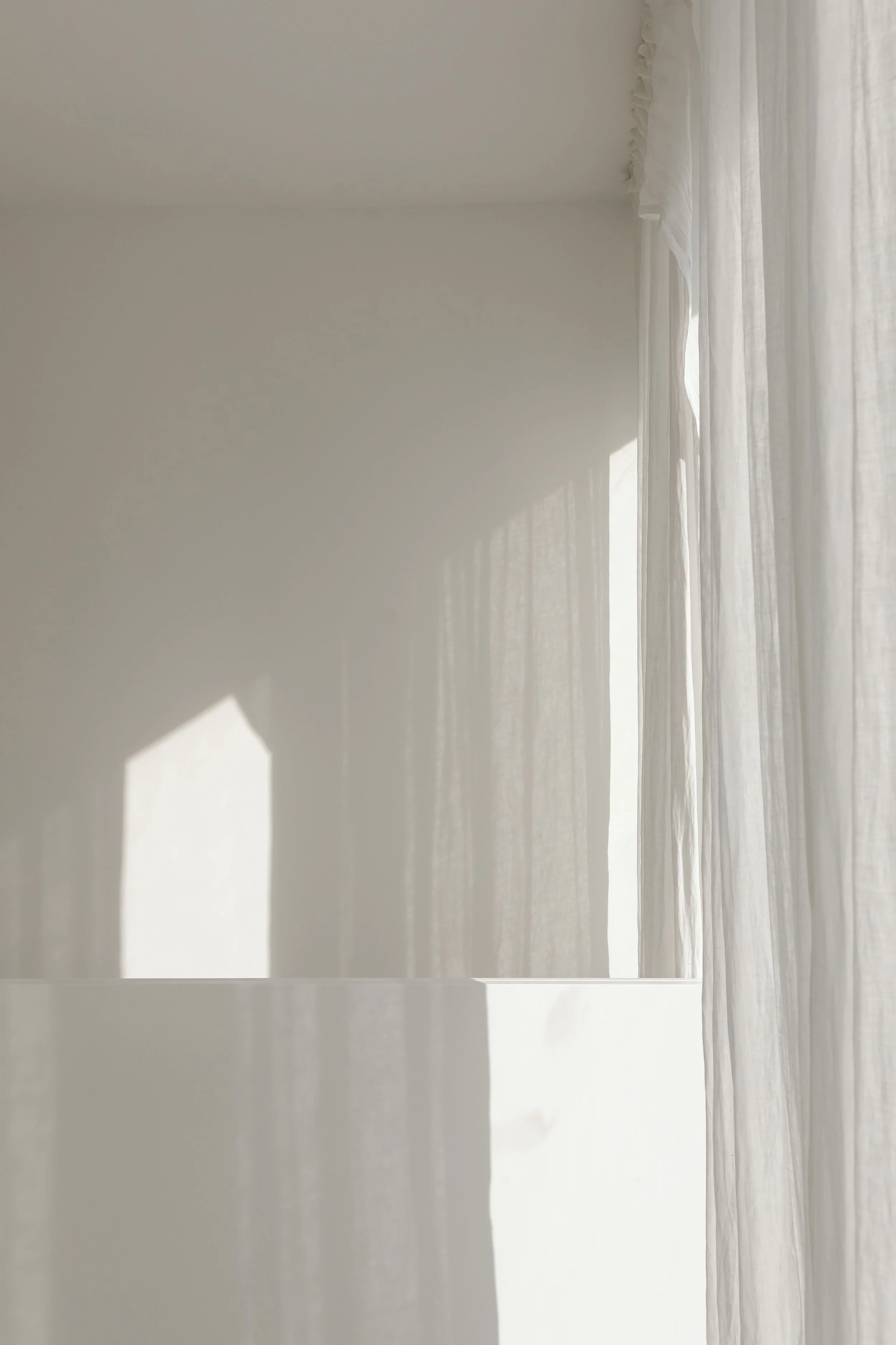 Sunlight shining through sheer white curtains onto a white wall and surface, creating shadows.