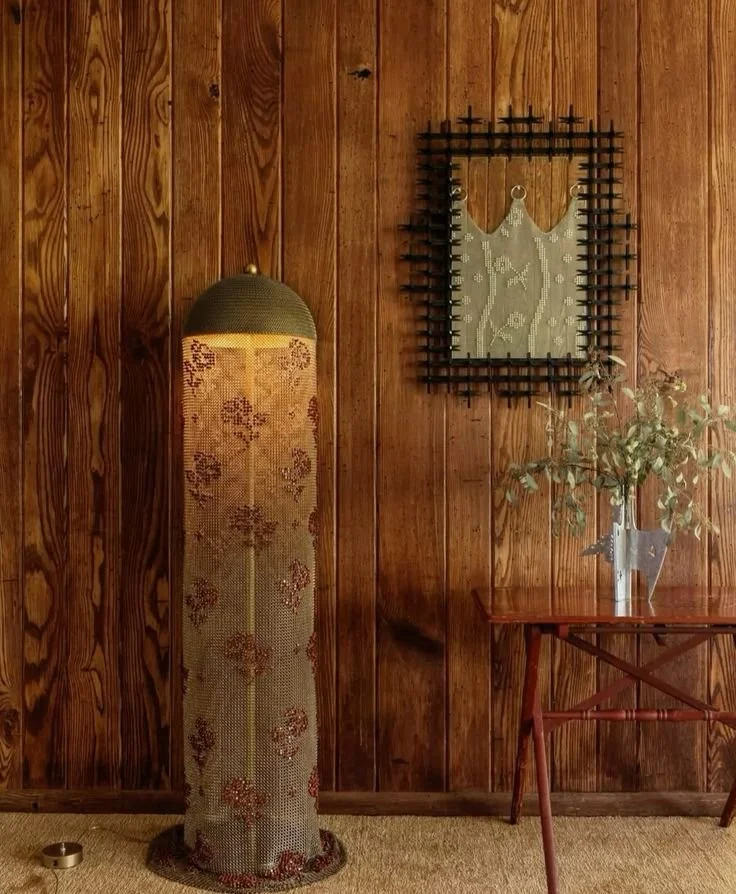 A vintage-style standing lamp with a brass base and fabric shade, standing next to a wooden table with a metal watering can filled with greenery, against a wooden paneled wall decorated with a framed piece of art with a lace-like pattern.