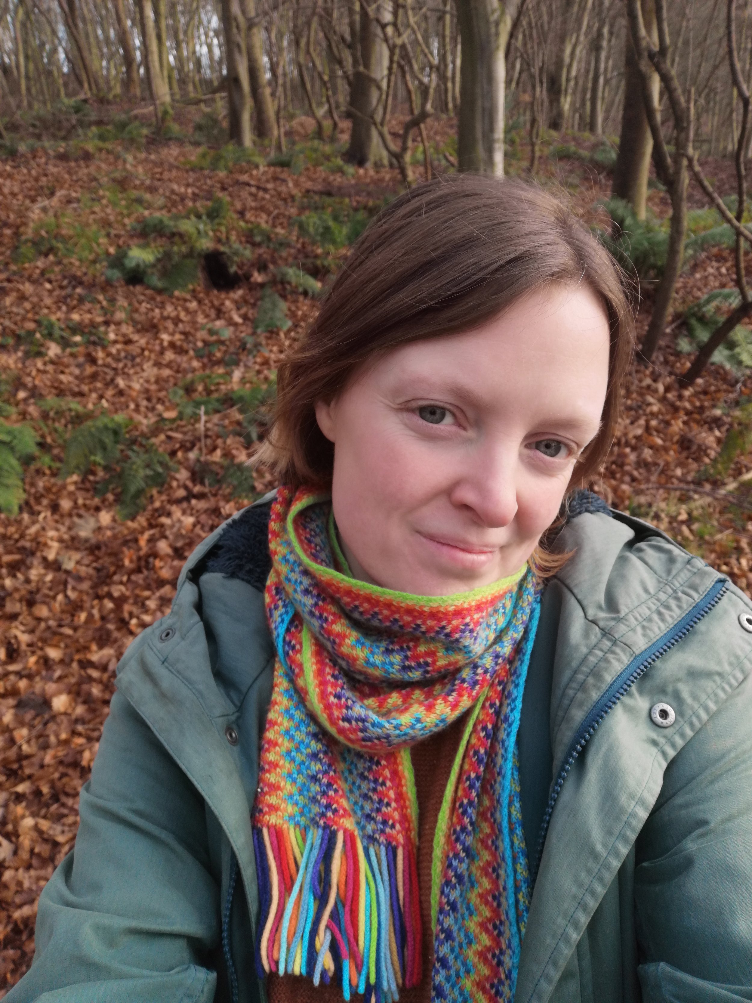 A woman taking a selfie outdoors in a wooded forest during autumn, wearing a colorful scarf and a green jacket.