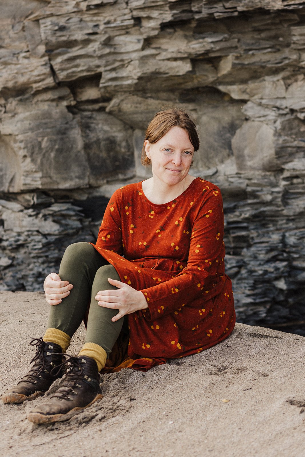 A woman sitting on sand near a rocky cliff, wearing a rust-colored dress and hiking boots.