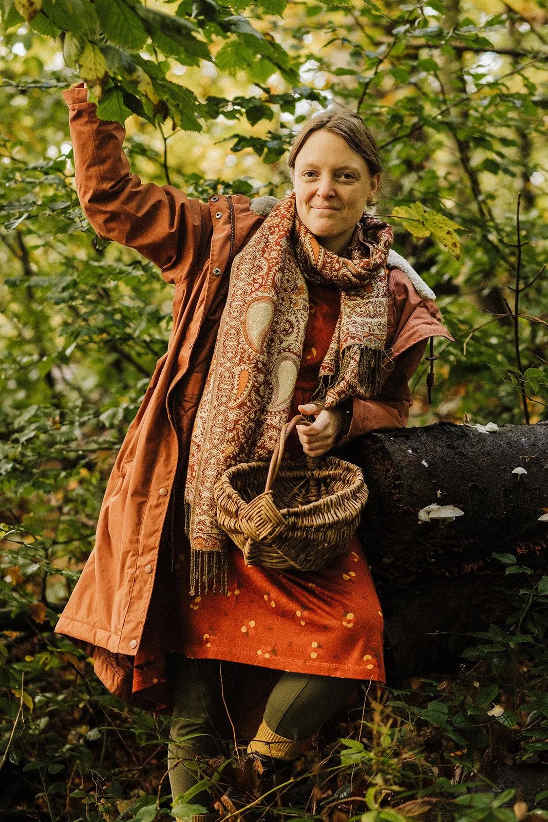 A woman wearing a brown coat and patterned scarf is holding a woven basket while standing outdoors in a leafy forest, leaning on a fallen tree.