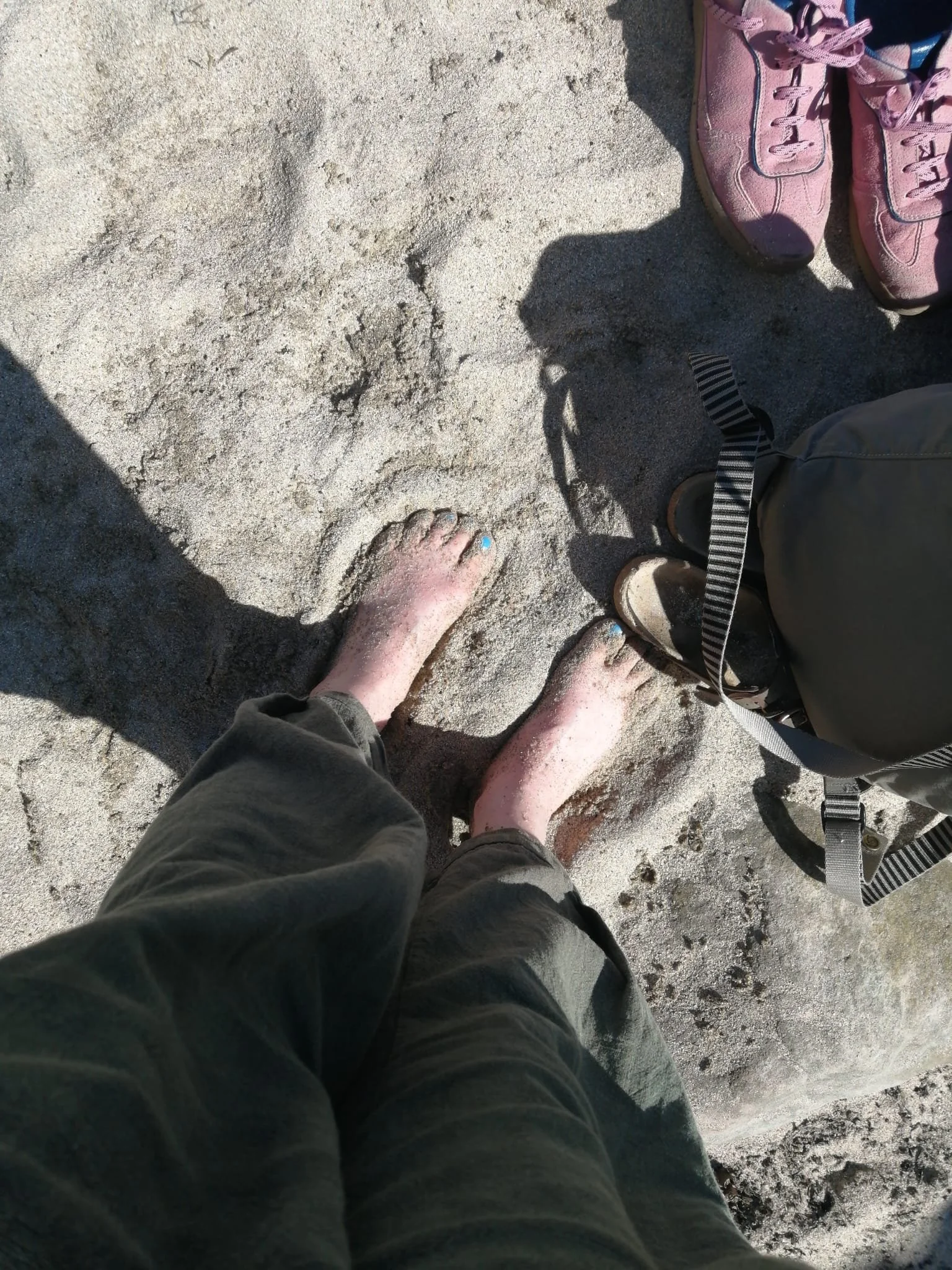 Person standing barefoot on sandy beach next to a backpack and pink sneakers.