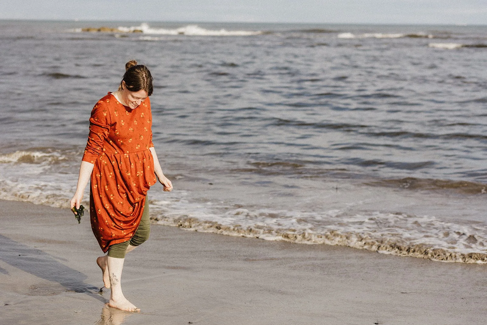 A woman wearing an orange dress walks barefoot along the shoreline of the beach, smiling and looking down at the sand, seaweed in hand.