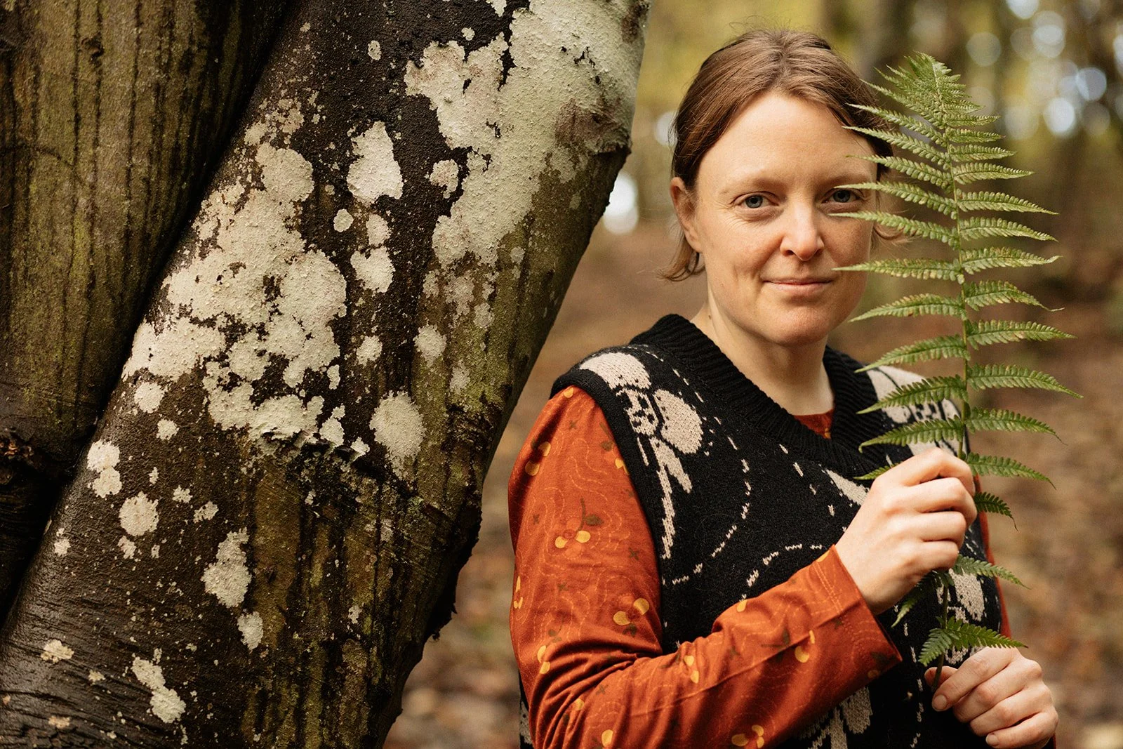 A woman with short brown hair holding a fern leaf outdoors in a wooded area.