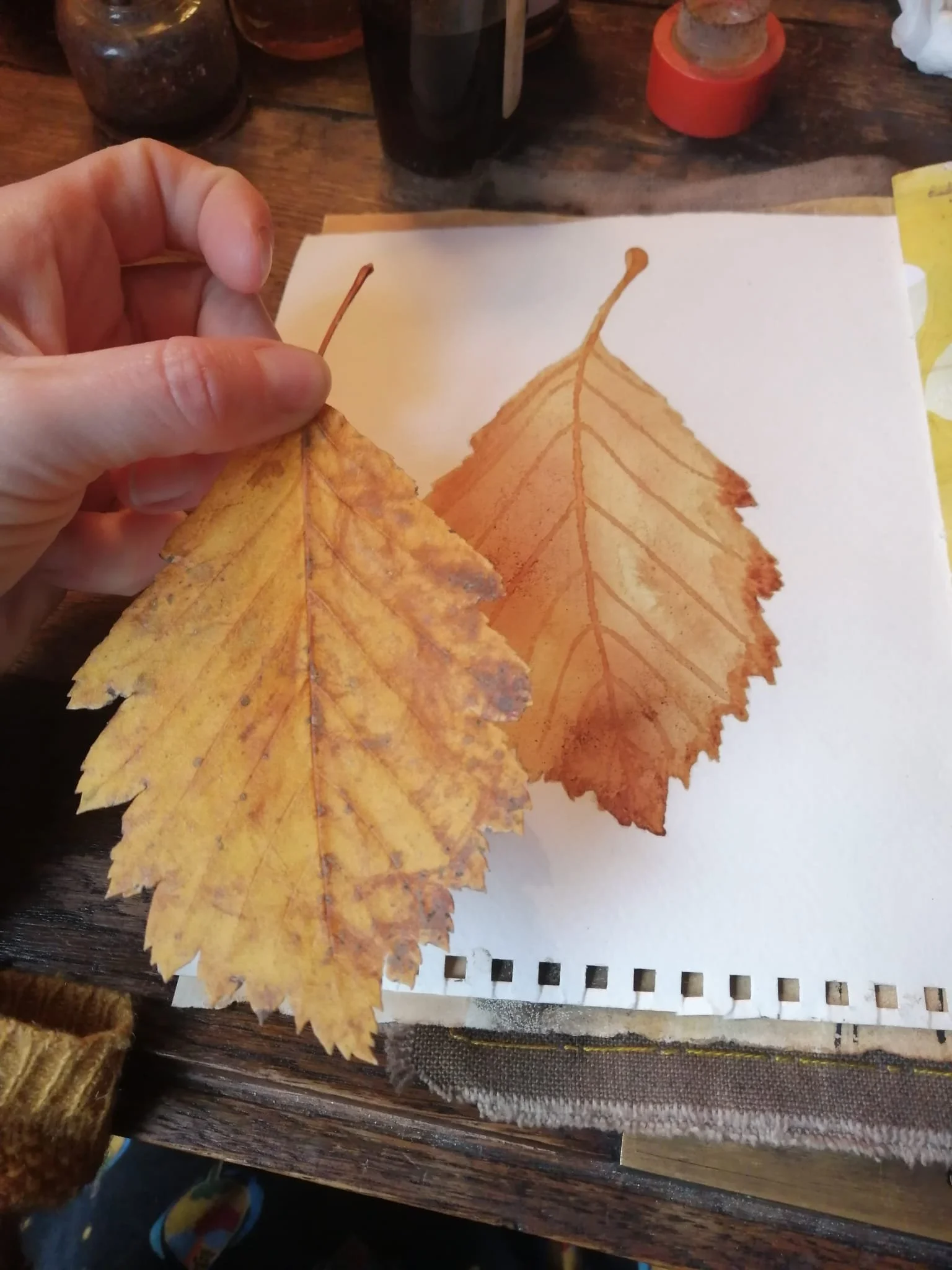 A person holding a yellow and brown autumn leaf, with a painted depiction of the same leaf on paper underneath, on a wooden table with art supplies in the background.