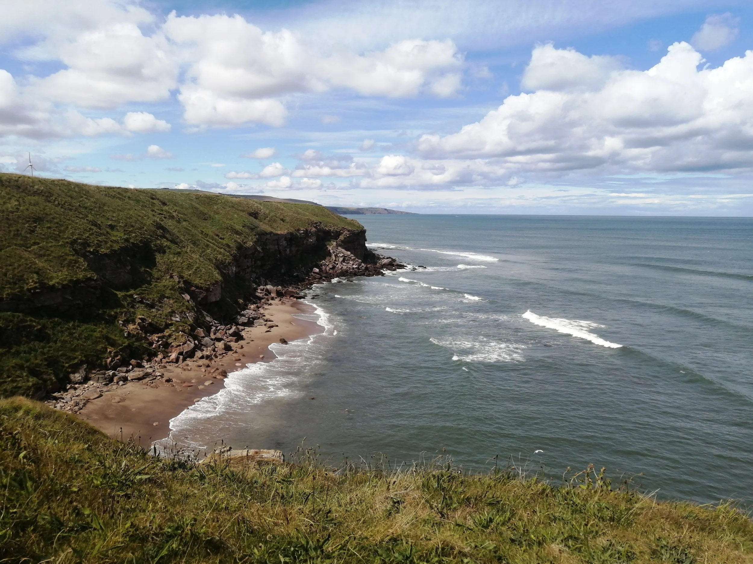 Clifftop view of a sandy beach with rocks, green grass on top, and ocean waves under a partly cloudy sky.