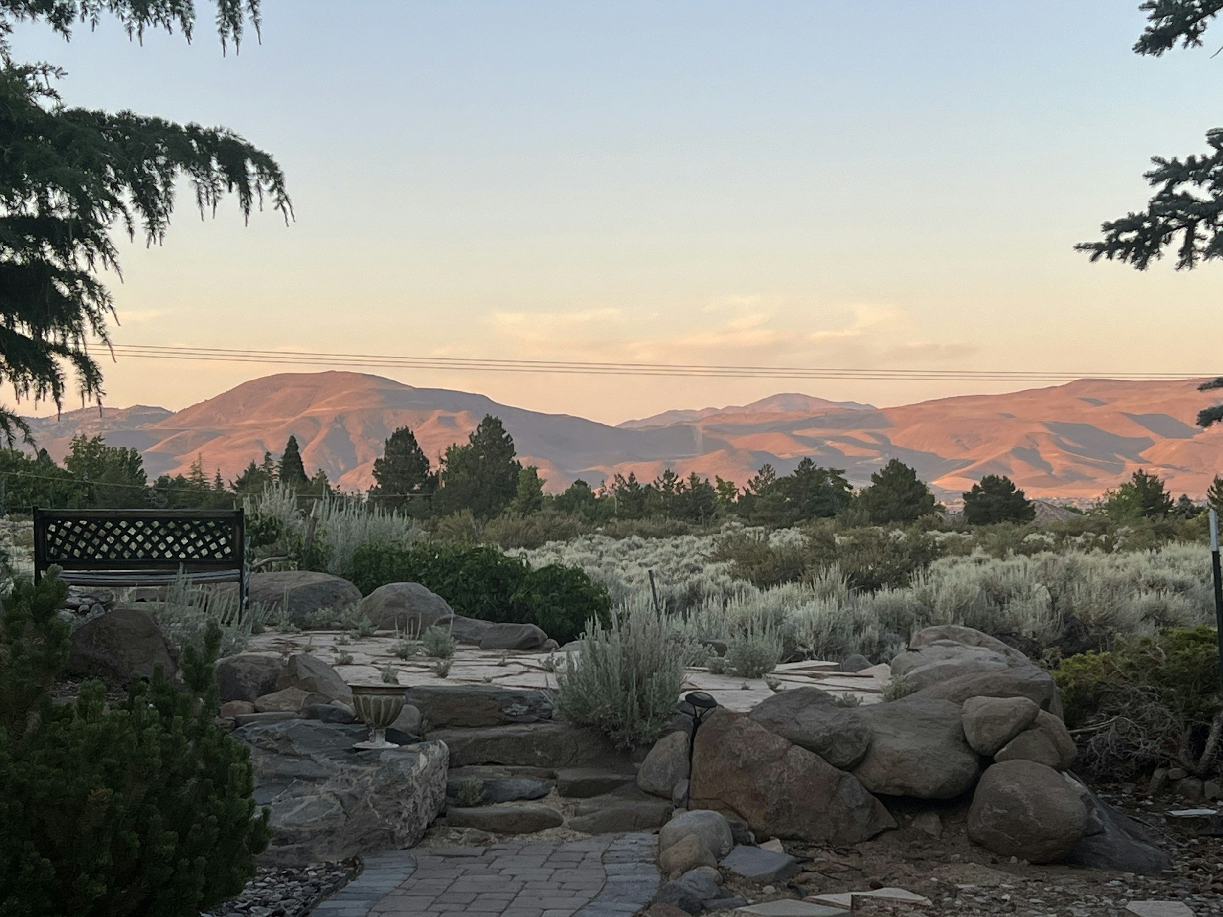 Scenic view of a desert landscape with mountains in the distance, trees, rocks, and a patio with a bench and decorative stone pathway in the foreground.