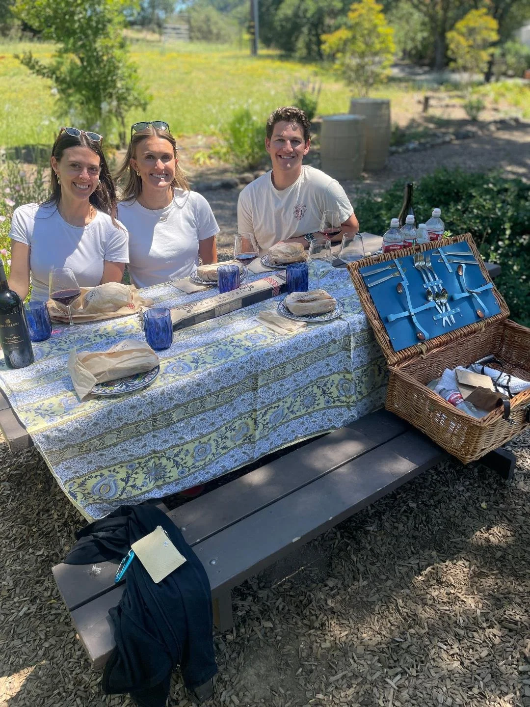 Three women and one man sitting at a picnic table outdoors with food and drinks, smiling.