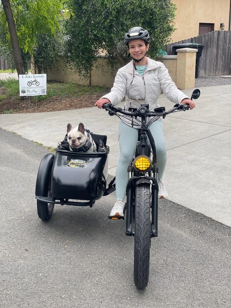 A woman wearing a bike helmet and light-colored athletic clothing riding an electric bike with a French bulldog in a front cargo bag. The dog looks happy with its tongue out. They are on a paved pathway with a sign for e-bike rentals in the background.