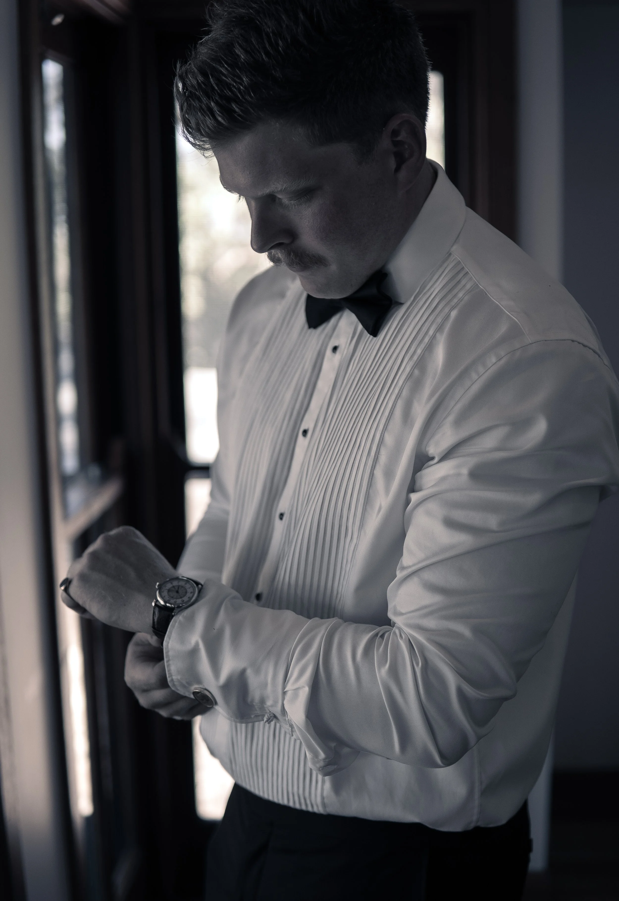 A man in a tuxedo and bow tie adjusting his watch inside a room with natural light coming through a window.