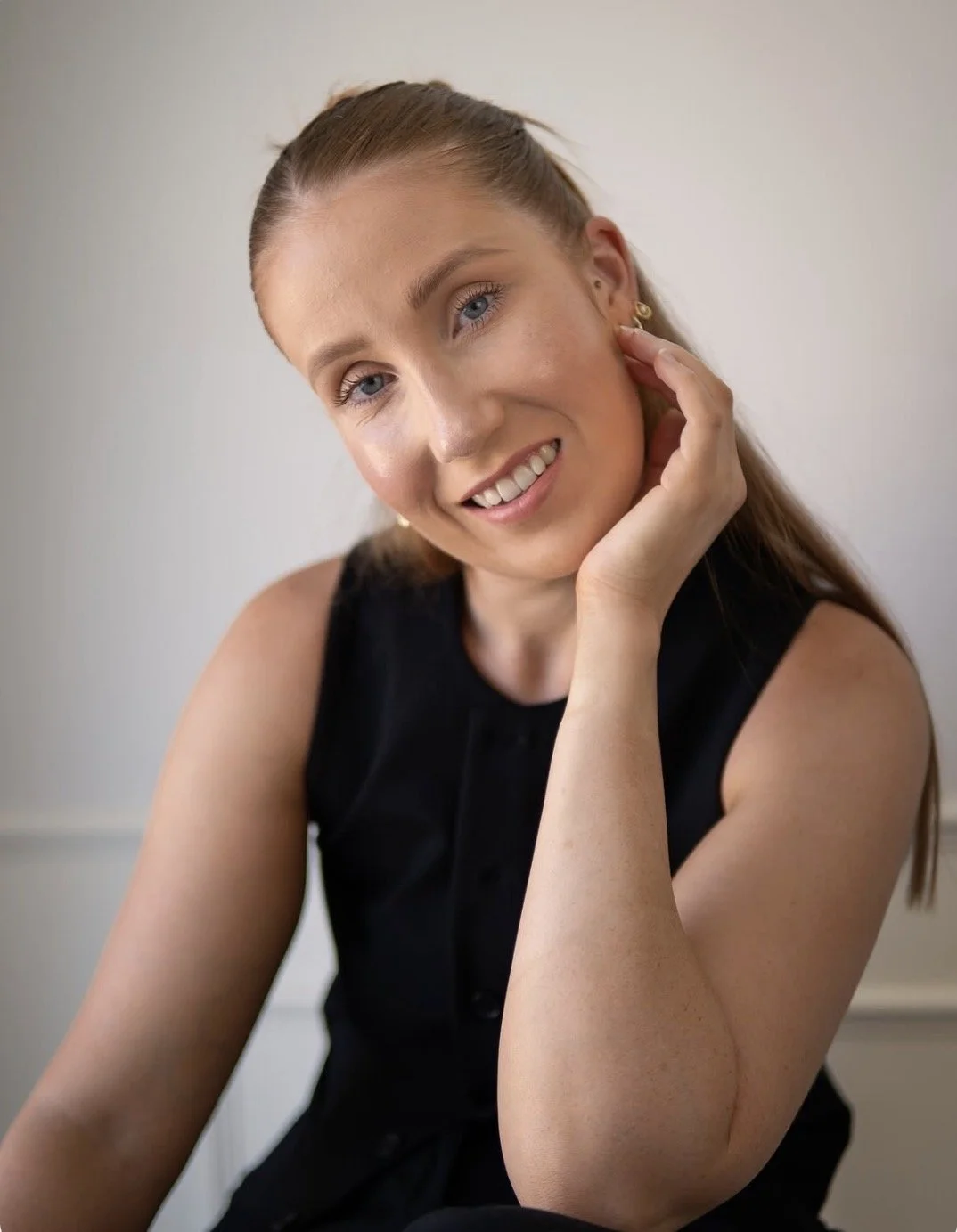 A young woman with blue eyes and light brown hair styled in a half-up half-down look, smiling gently and resting her chin on her hand, wearing a sleeveless black top and gold earrings, against a plain light-colored background.