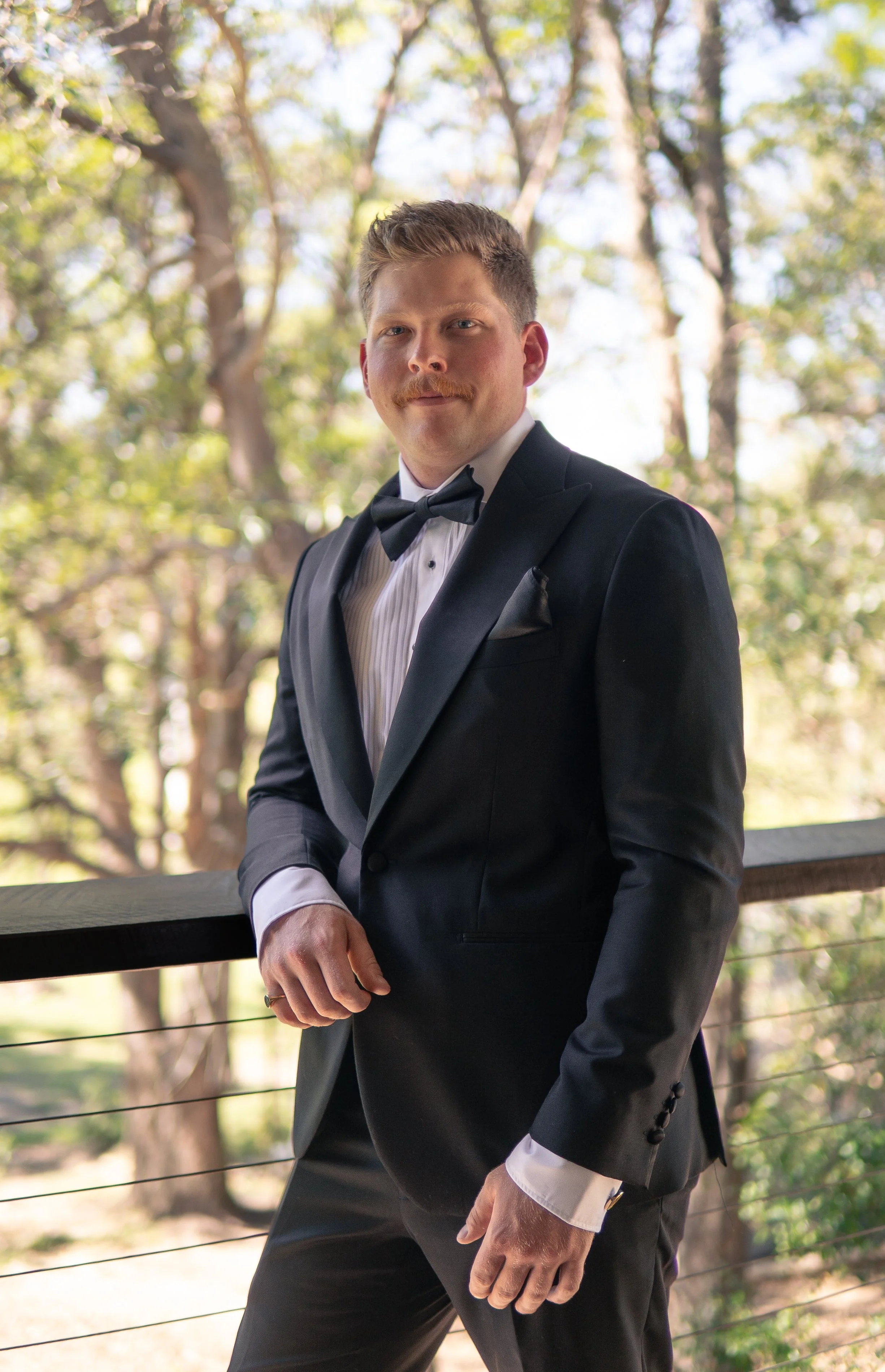 A man in a black tuxedo with a bow tie standing outdoors on a balcony with trees in the background.