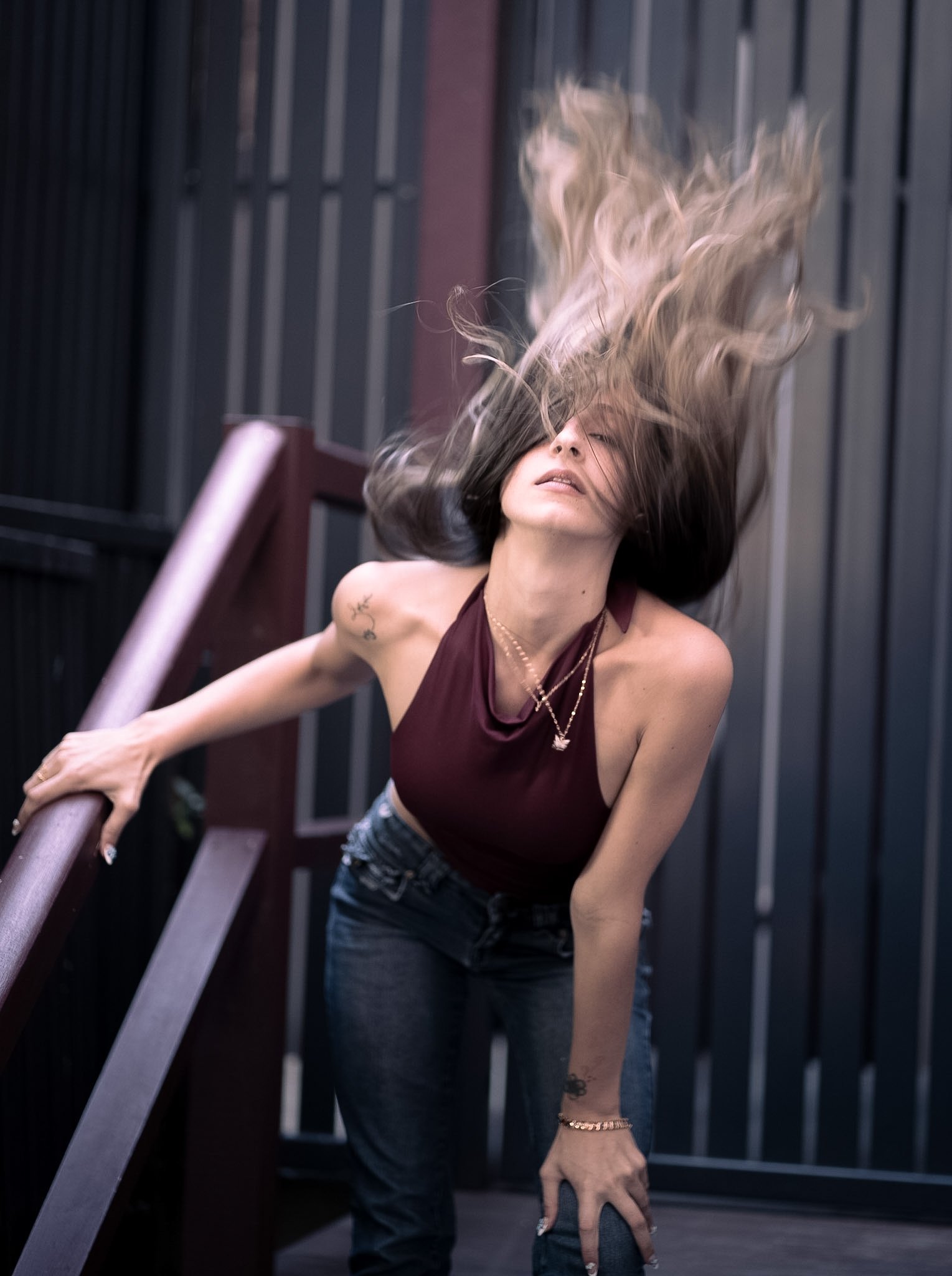 A woman with long hair flipping her hair while leaning on a wooden railing on an outdoor balcony or staircase.