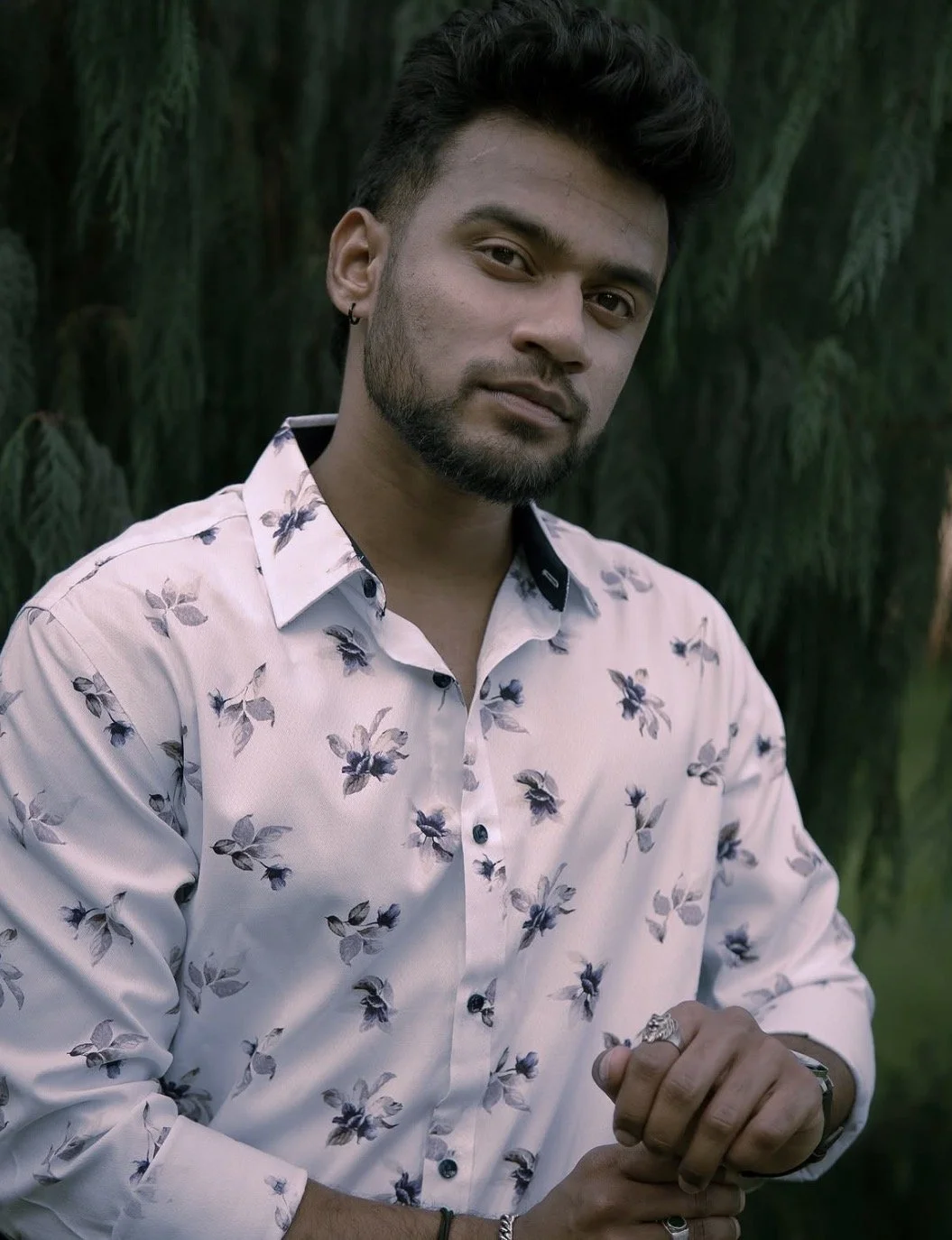 A young man with dark hair and a beard, wearing a floral-patterned white shirt, standing outdoors near a pine tree with a serious expression.