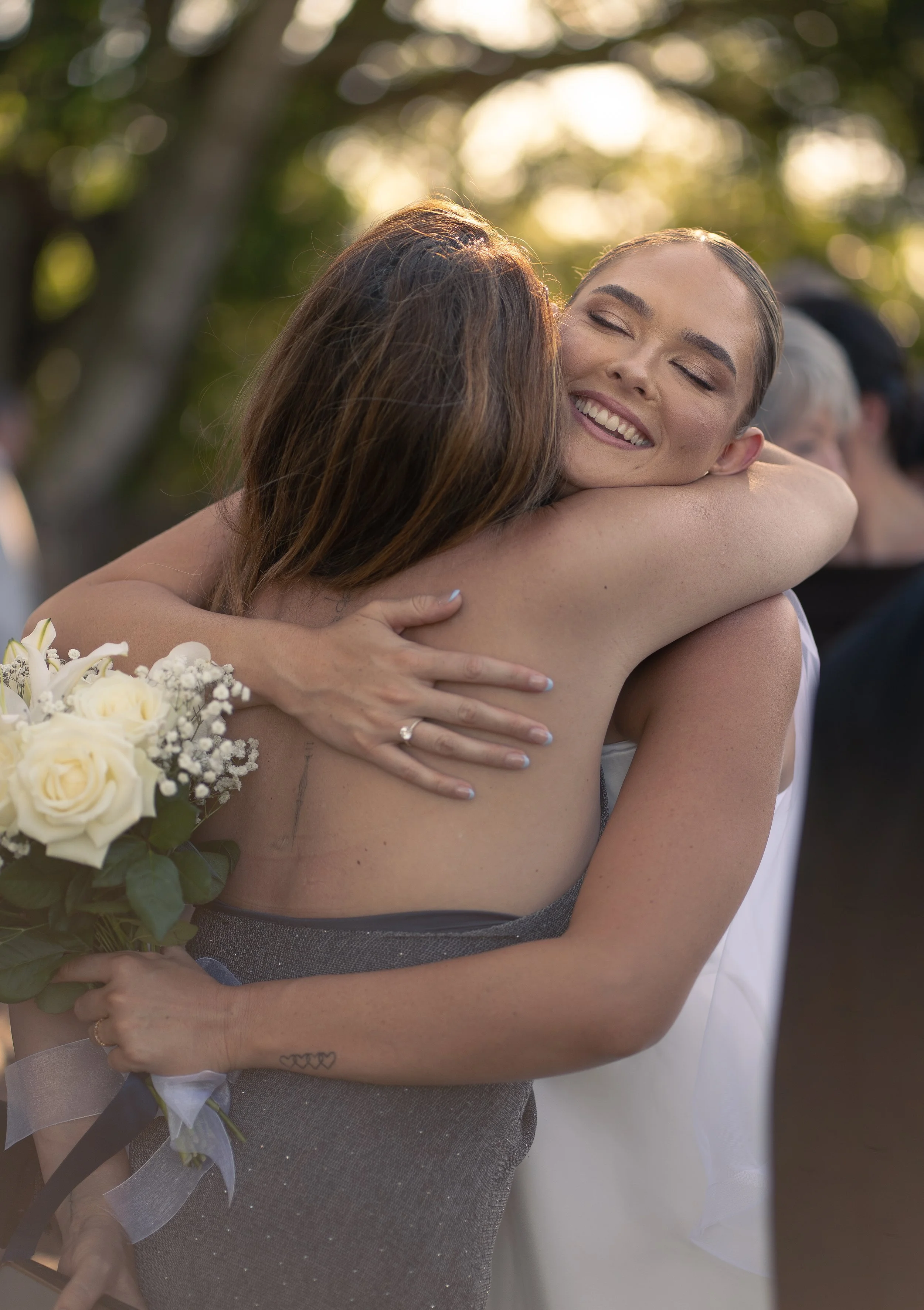 Two women hugging, one with dark hair and the other with light brown hair, during a wedding celebration outdoors. The woman with dark hair is holding a bouquet of white roses and has a small tattoo on her arm. The woman with light brown hair is smiling with her eyes closed.