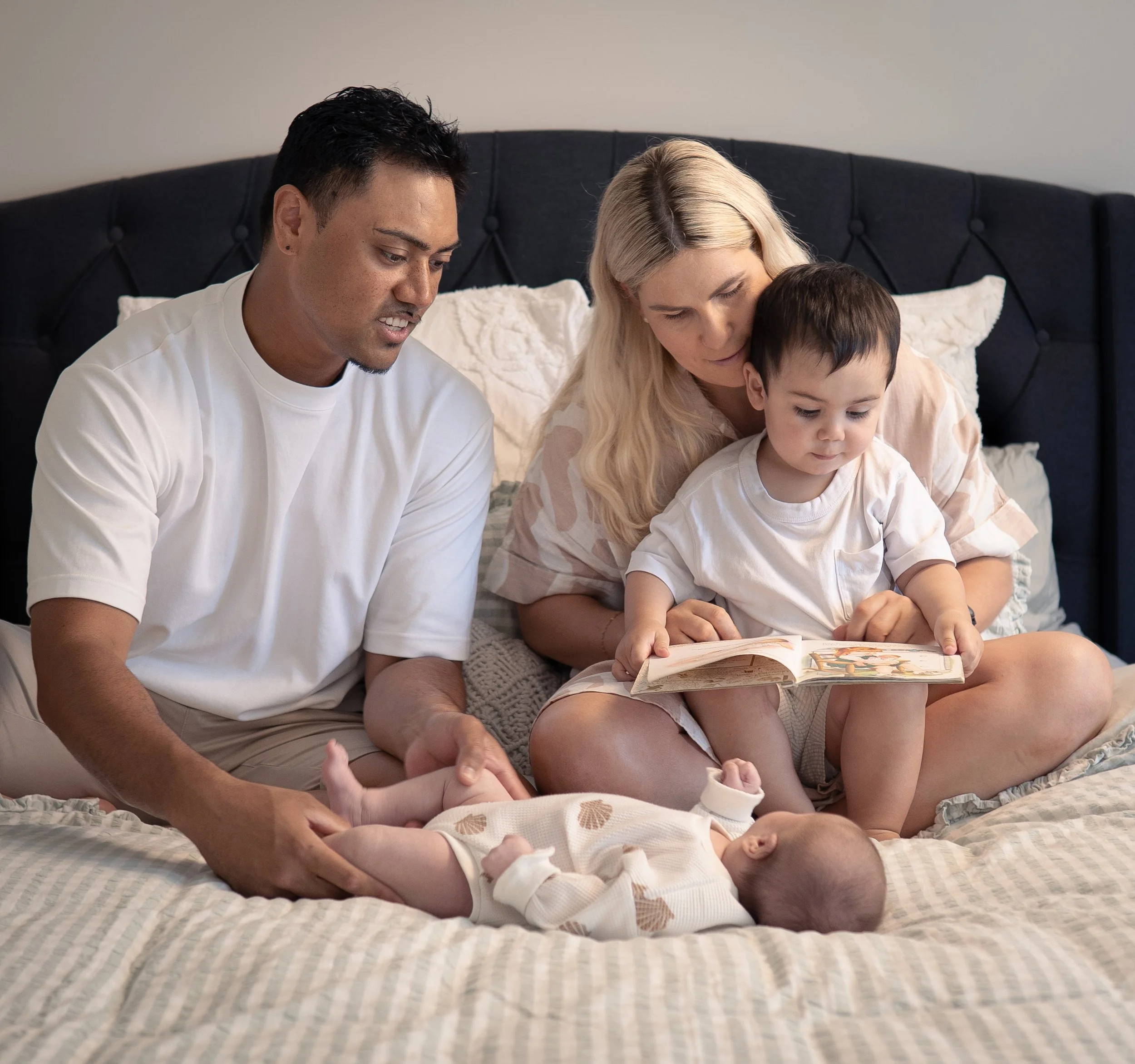 A family of four sitting on a bed, with a man holding a baby, a woman and a young boy looking at a book together, all in a bedroom.