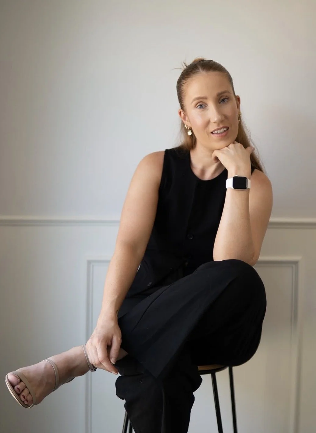 Woman with long red hair, wearing a black sleeveless top and black pants, sitting on a chair, resting her chin on her hand, and looking at the camera. She is wearing earrings and a smartwatch on her left wrist, with a neutral background and white wall paneling.