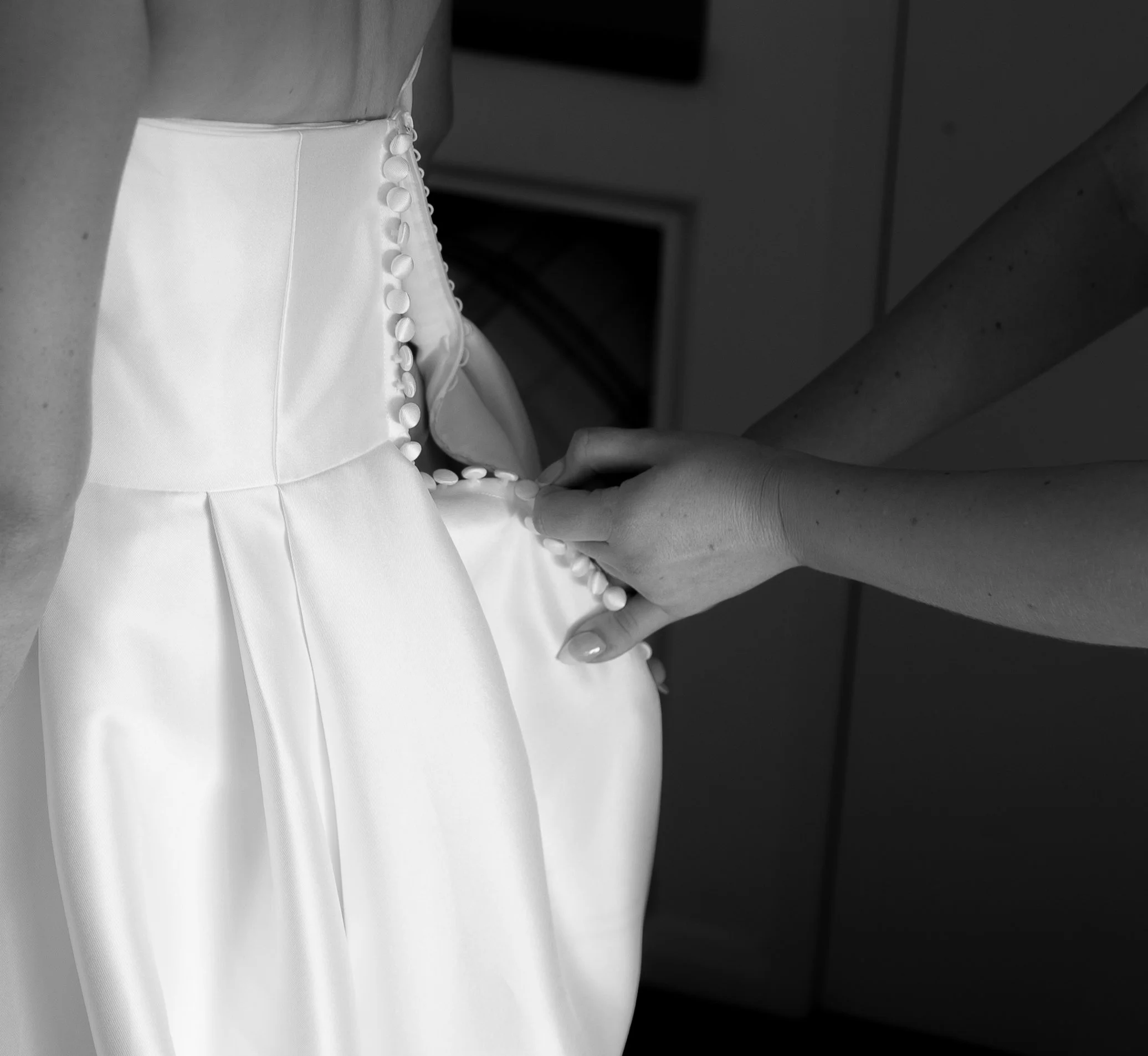 A person is buttoning the back of a white satin wedding dress, with another person's hands helping.