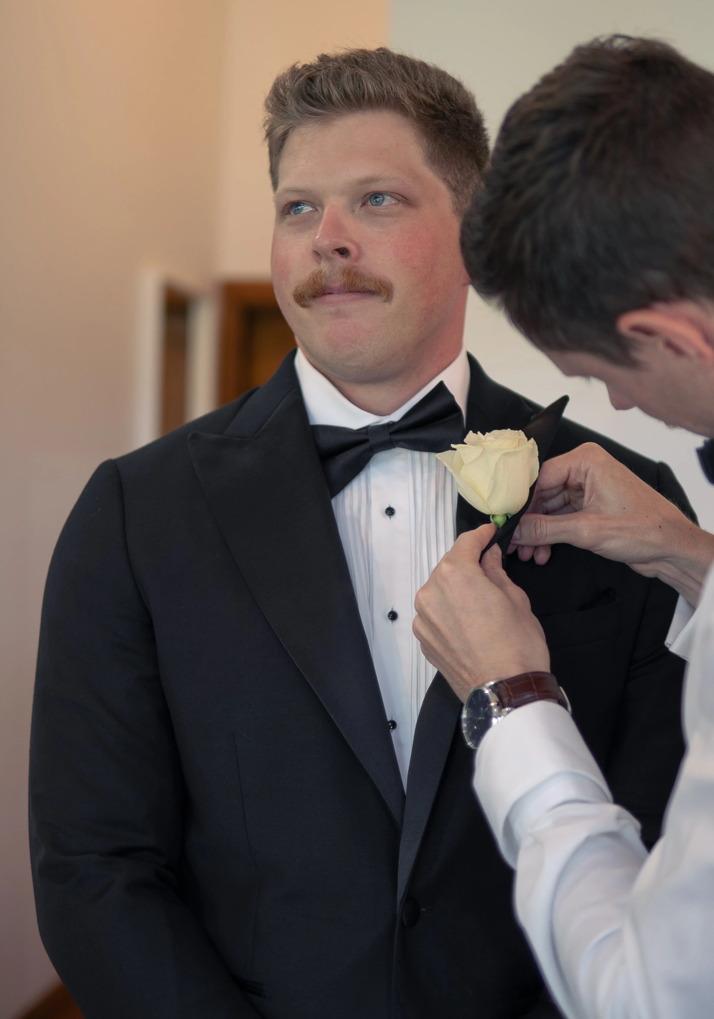 A groom with a mustache wearing a tuxedo with a bow tie is having a white rose pinned to his lapel by another man during a wedding.