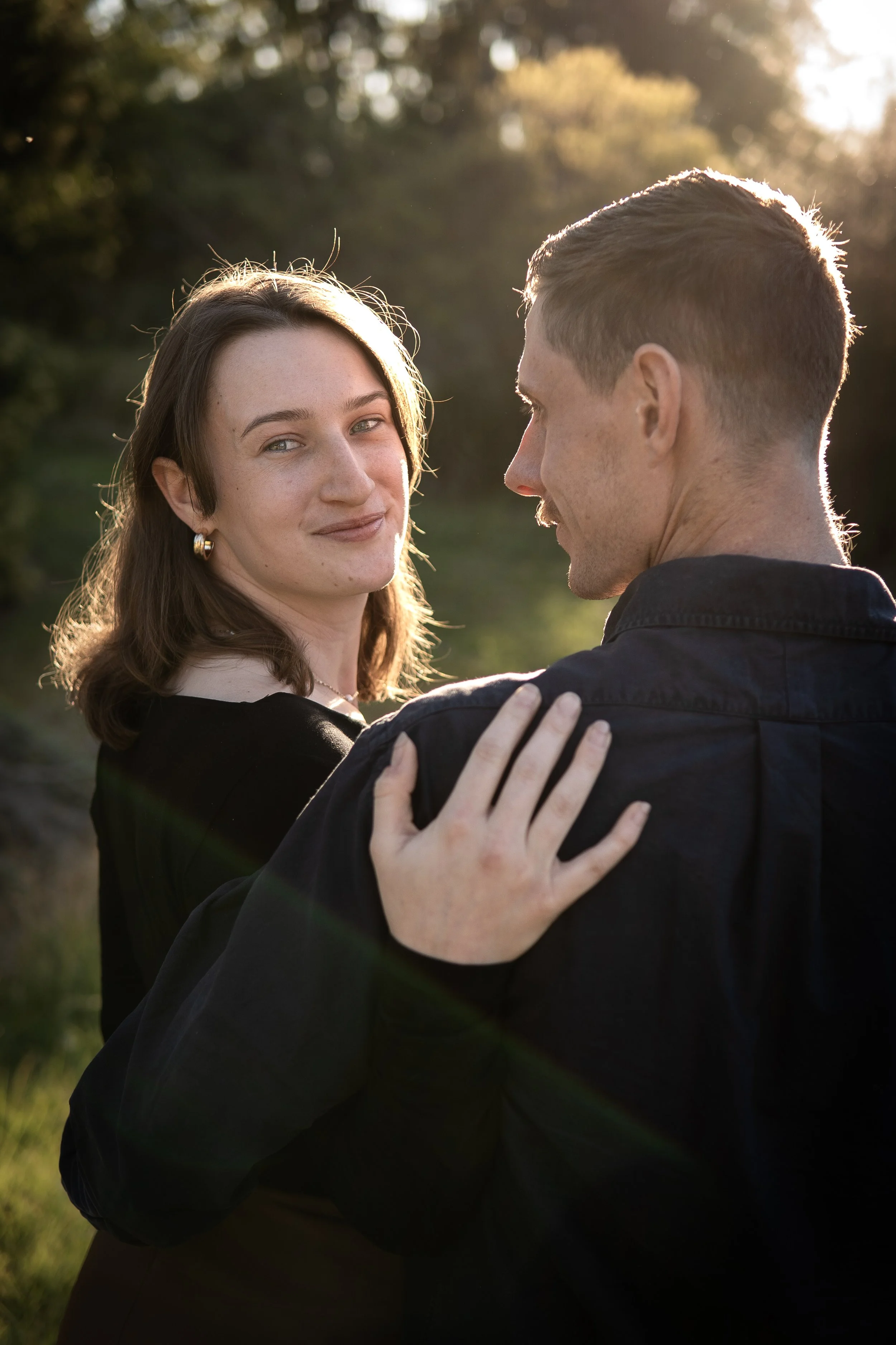 A young woman with shoulder-length brown hair and earrings looks at the camera, while a man with short hair faces her and holds her shoulder in an outdoor setting during golden hour.