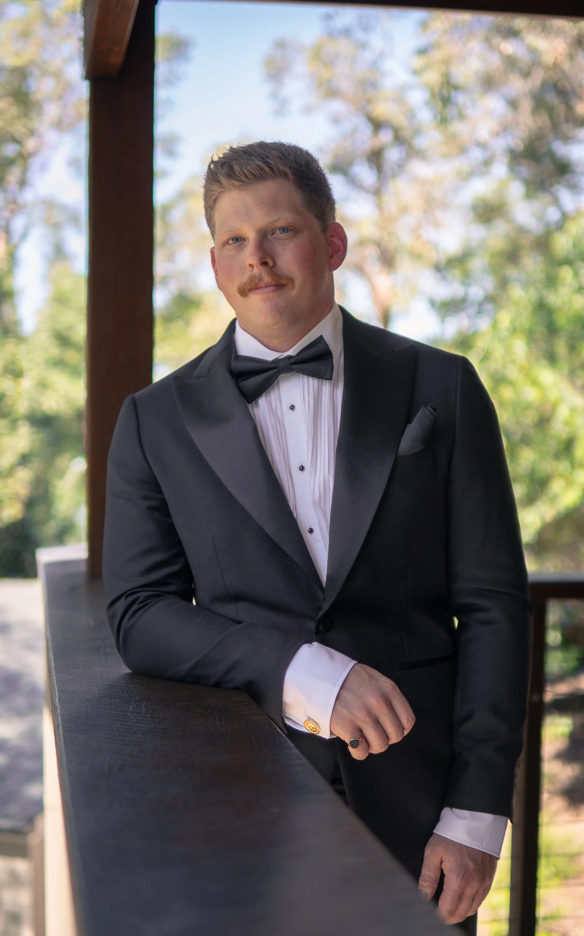 A man in a tuxedo with a bow tie, standing outdoors on a balcony with trees in the background.