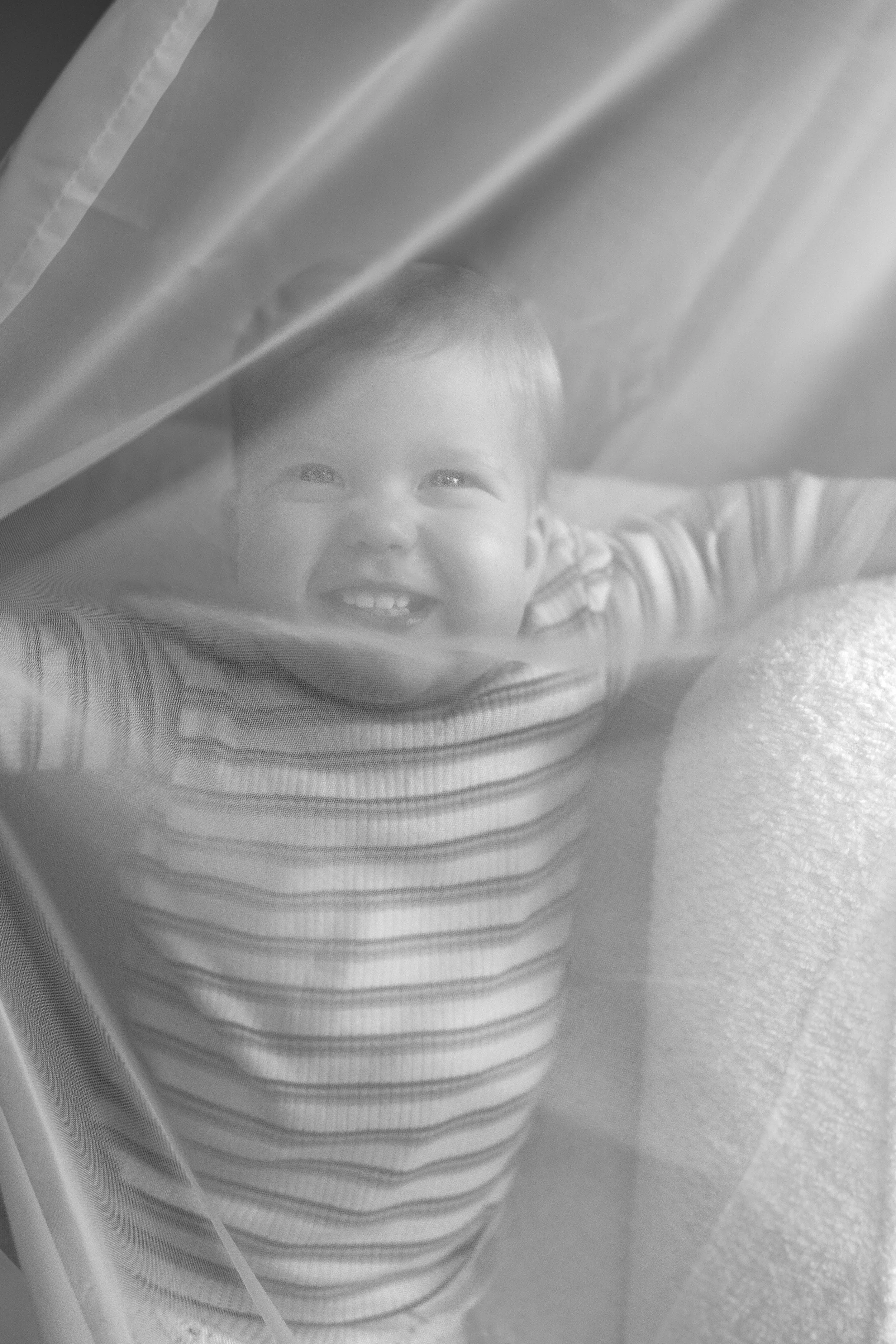 A young child with a big smile, seen through a glass window, with a curtain partially covering the view.
