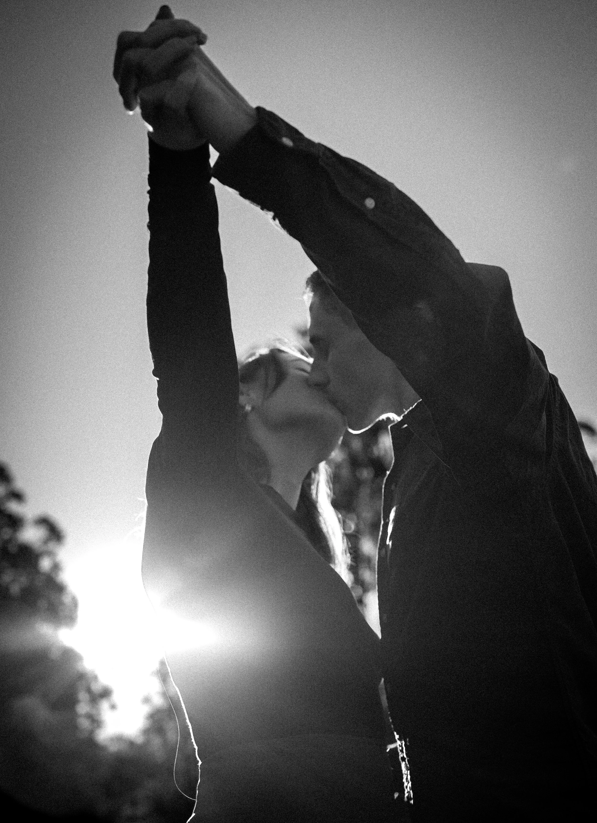 Black and white photo of a couple kissing outdoors, with the sun setting in the background.