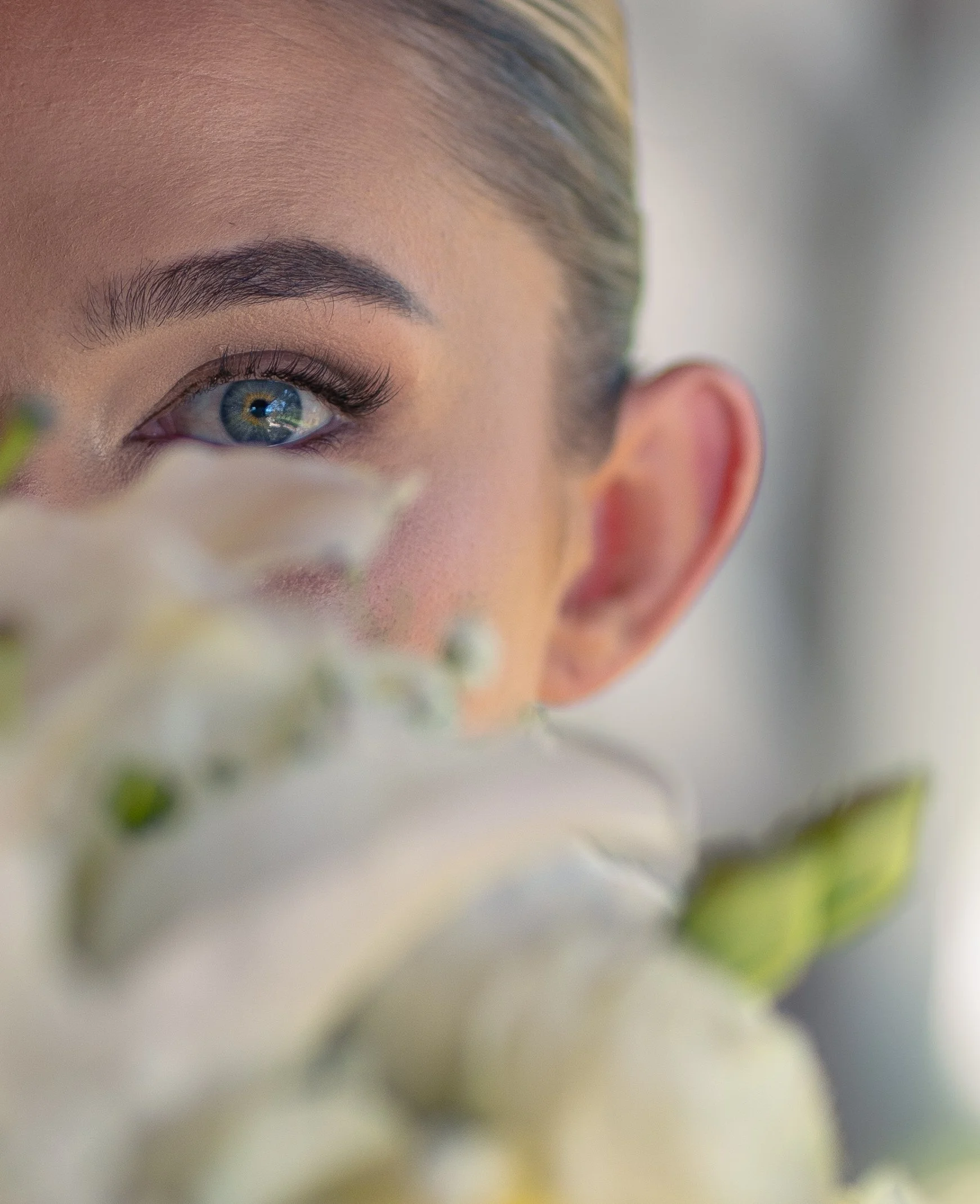 Close-up of a woman with blue eyes, blonde hair, and well-groomed eyebrows, partially hidden behind white lilies.