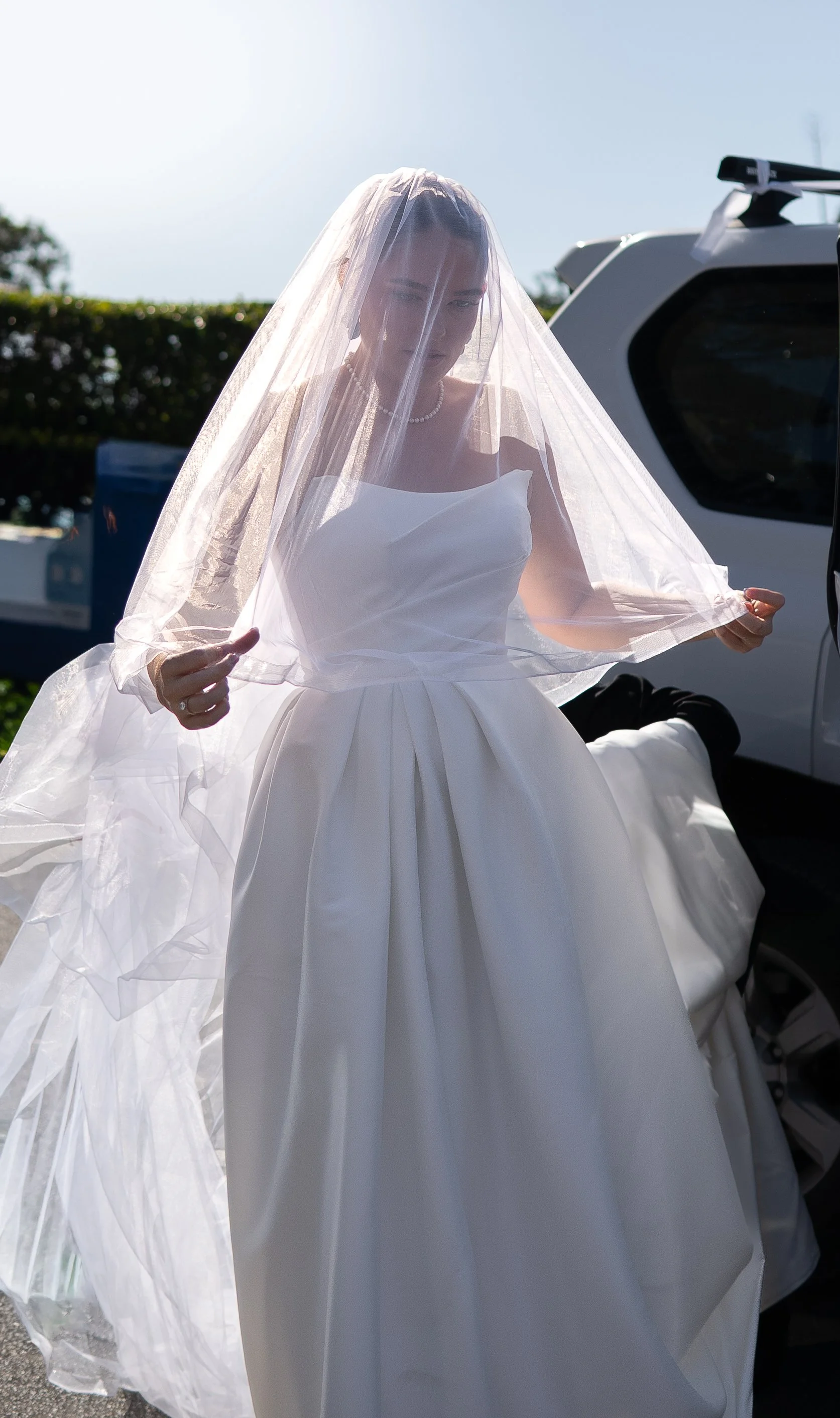 Bride in a white wedding dress and veil standing outdoors, holding the veil in her hands, with a vehicle and trees in the background.