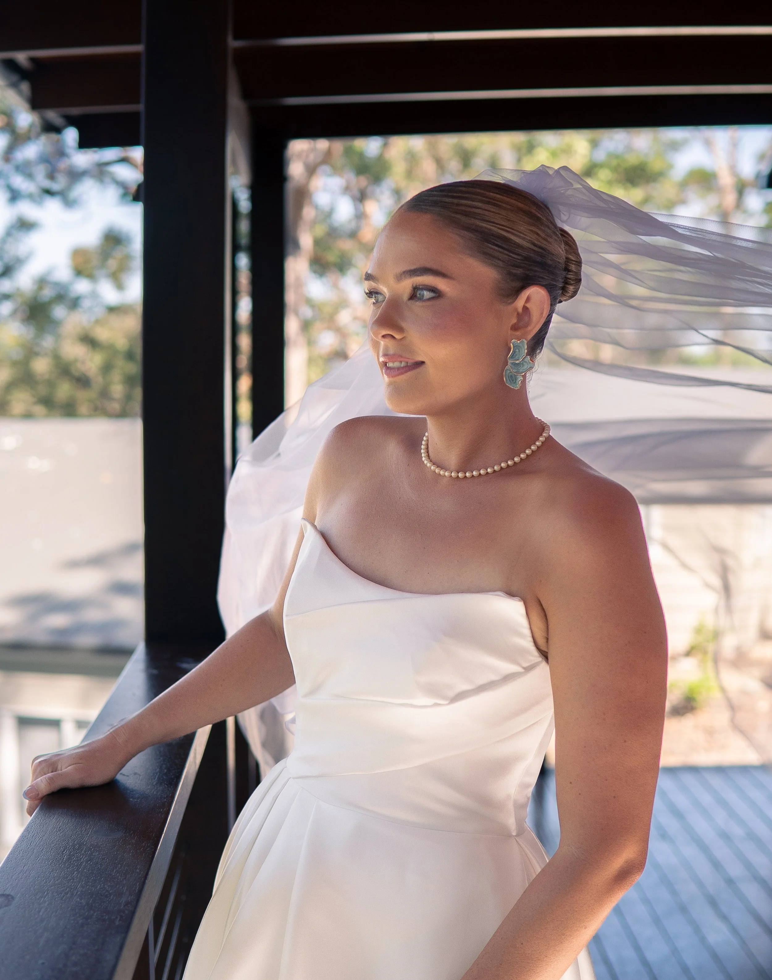 Bride in a white wedding dress standing on a balcony, looking to the side with sunlight on her face and veil flowing behind her.