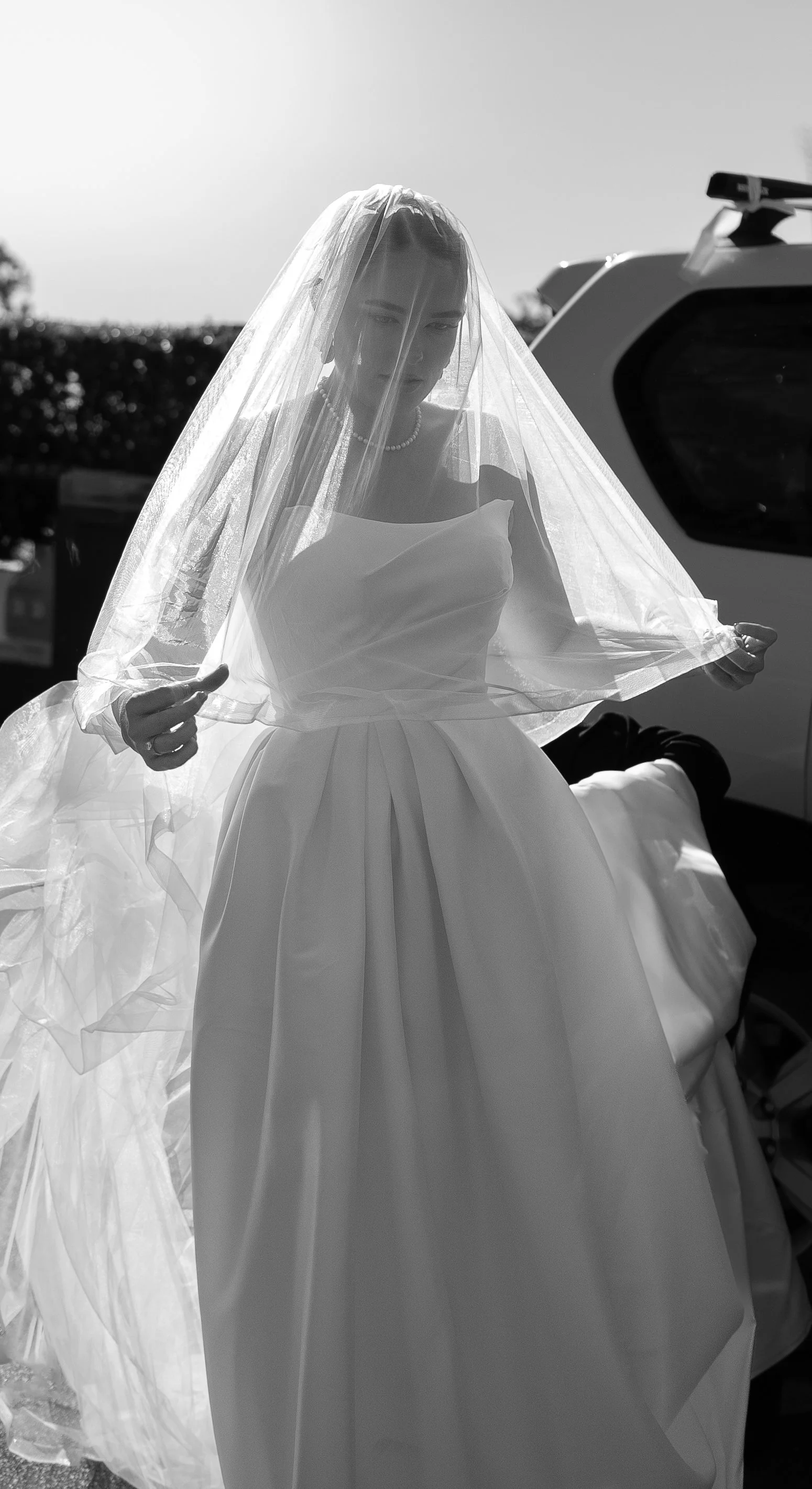 A woman in a wedding dress holding a veil over her head, standing outside near a vehicle.