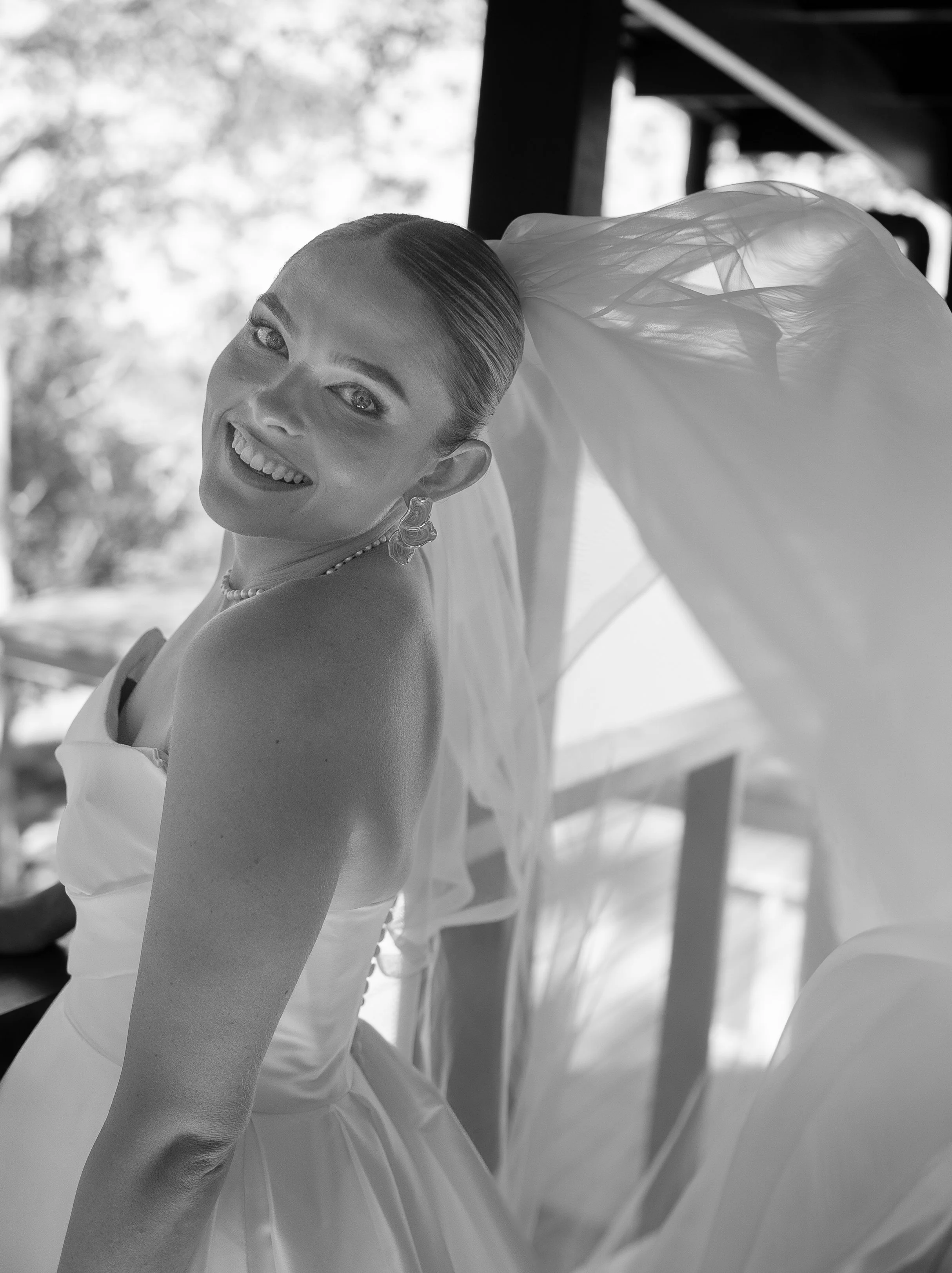 Black and white photo of a smiling woman in a wedding dress, with a veil flowing behind her, looking over her shoulder.