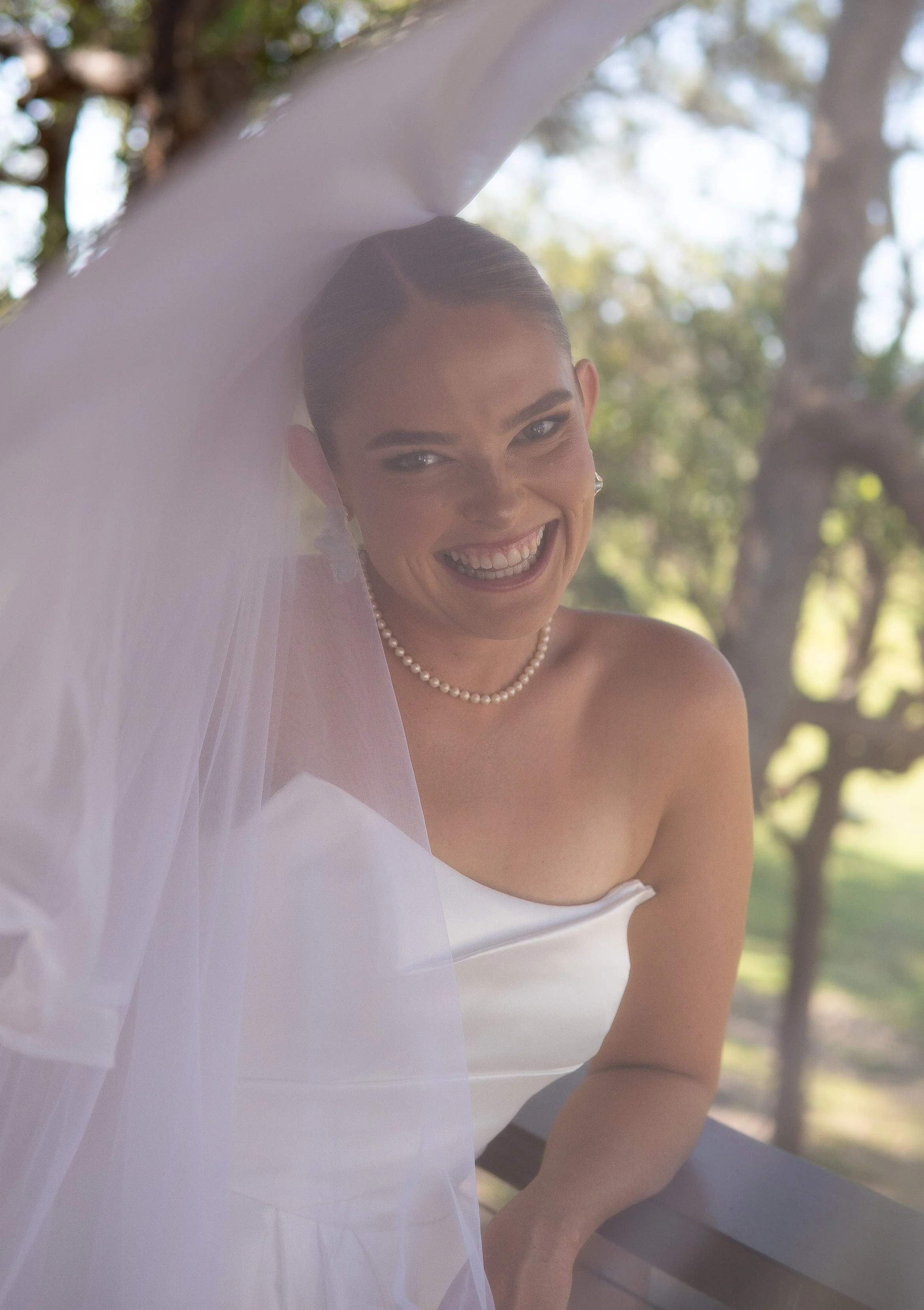 A smiling bride in a white wedding dress and pearl necklace, partially covered by a veil, leaning on a wooden railing outdoors with trees in the background.